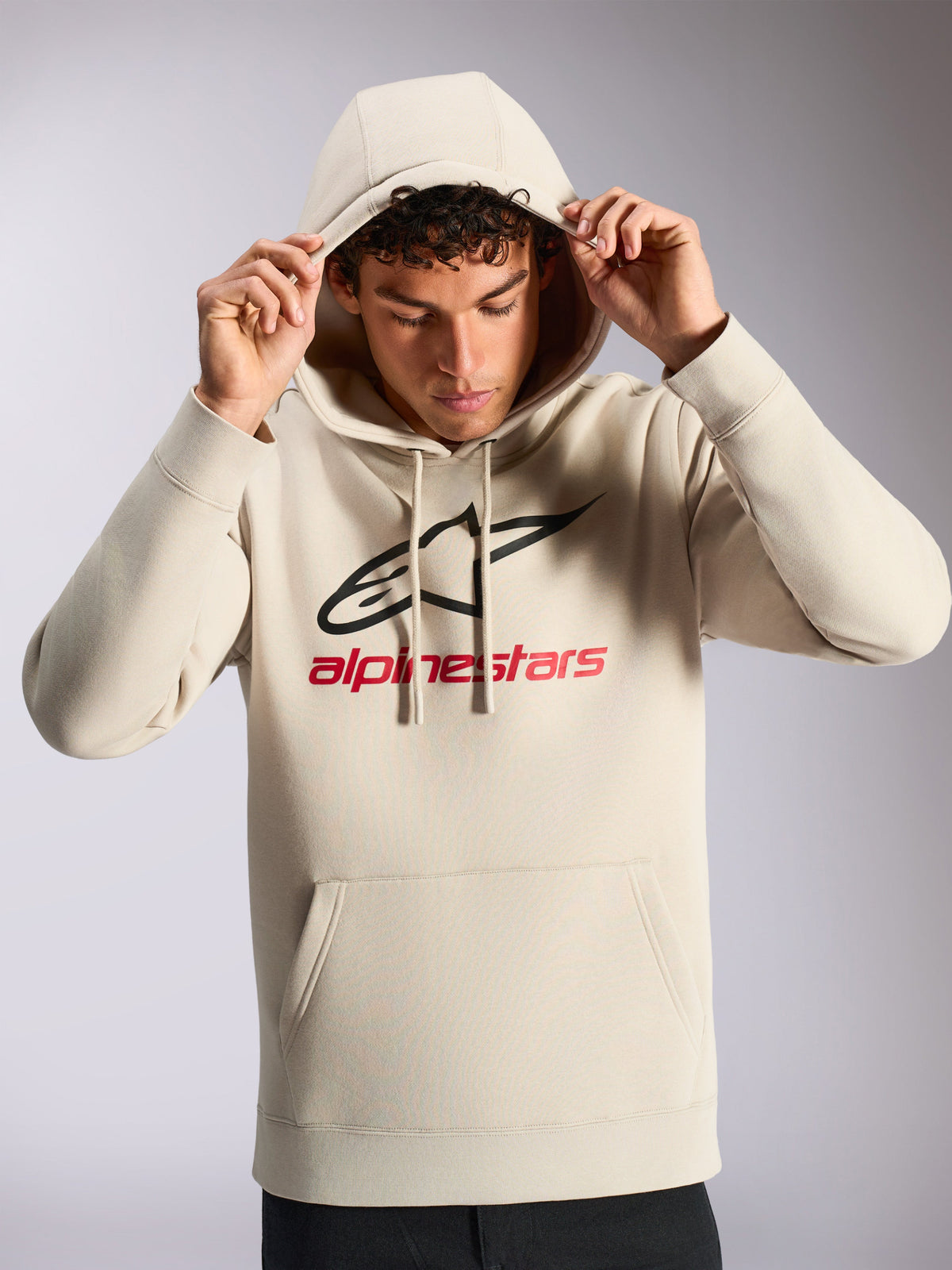 Alpinestars Always V3 Hoodie, modelo masculino con sudadera cerrada de forro polar en blanco roto, logotipo lineal delantero en negro y rojo, capucha con cordón, bolsillo tipo canguro, ropa de calle casual inspirada en el automovilismo