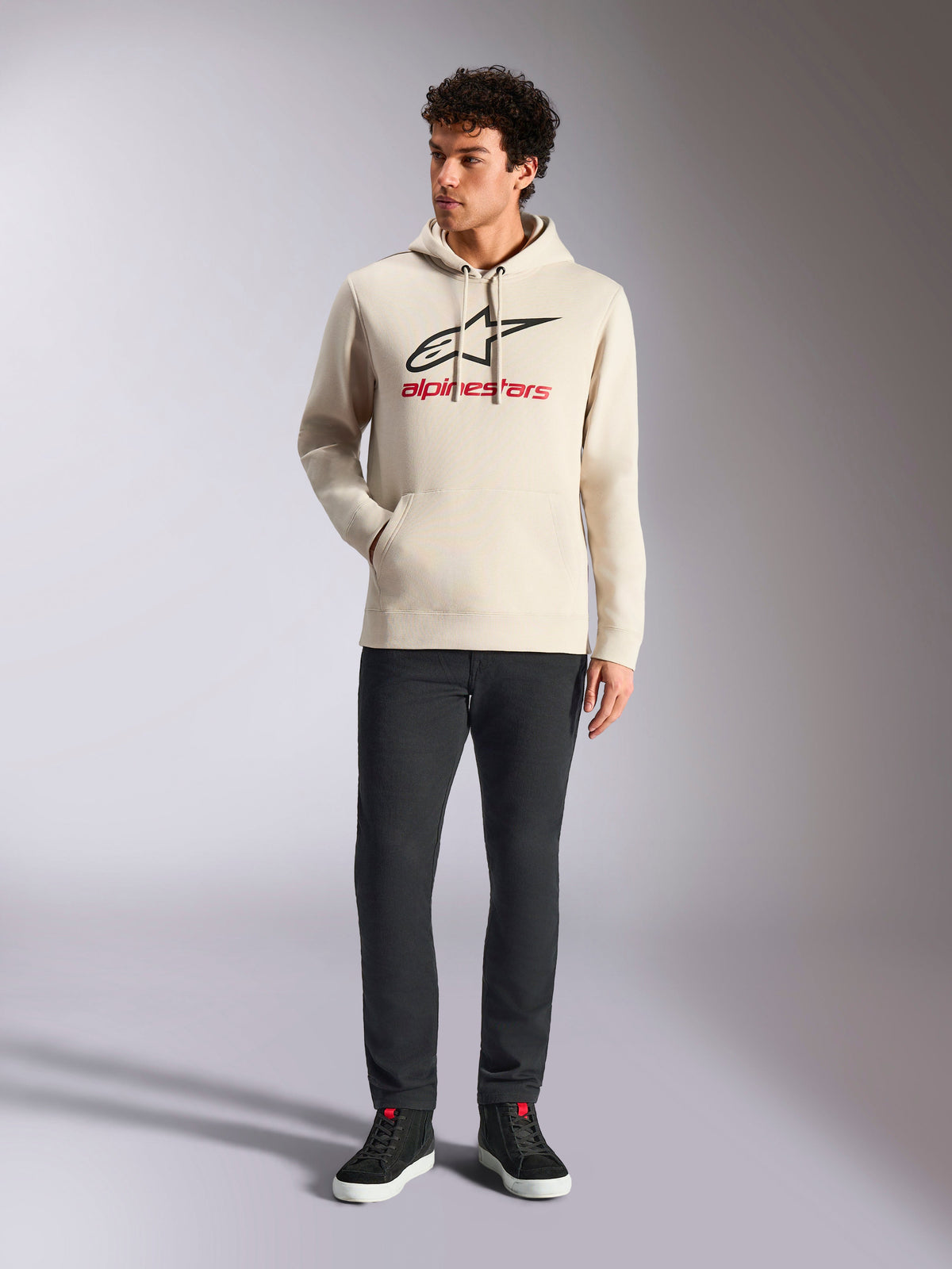 Sudadera Alpinestars Always V3, estilo streetwear informal, color blanco roto con logotipo de estrella negro y letras rojas, capucha con cordón, manga larga, usada por un modelo masculino con pantalones negros y zapatillas negras
