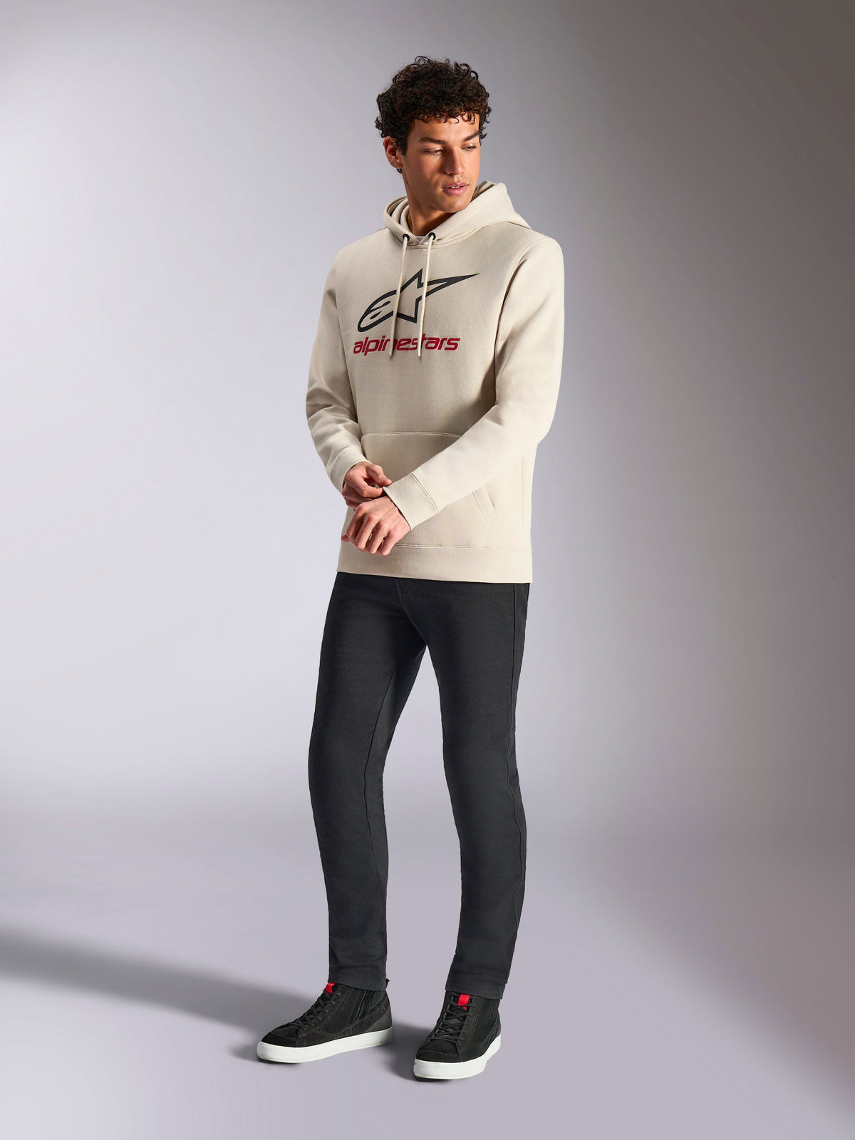 Alpinestars Always V3 Hoodie, sudadera de felpa en color blanco roto, logotipo lineal frontal con estrella negra y marca en rojo, lucida por un modelo masculino con pantalones negros y zapatillas negras, estilo casual motorsport streetwear