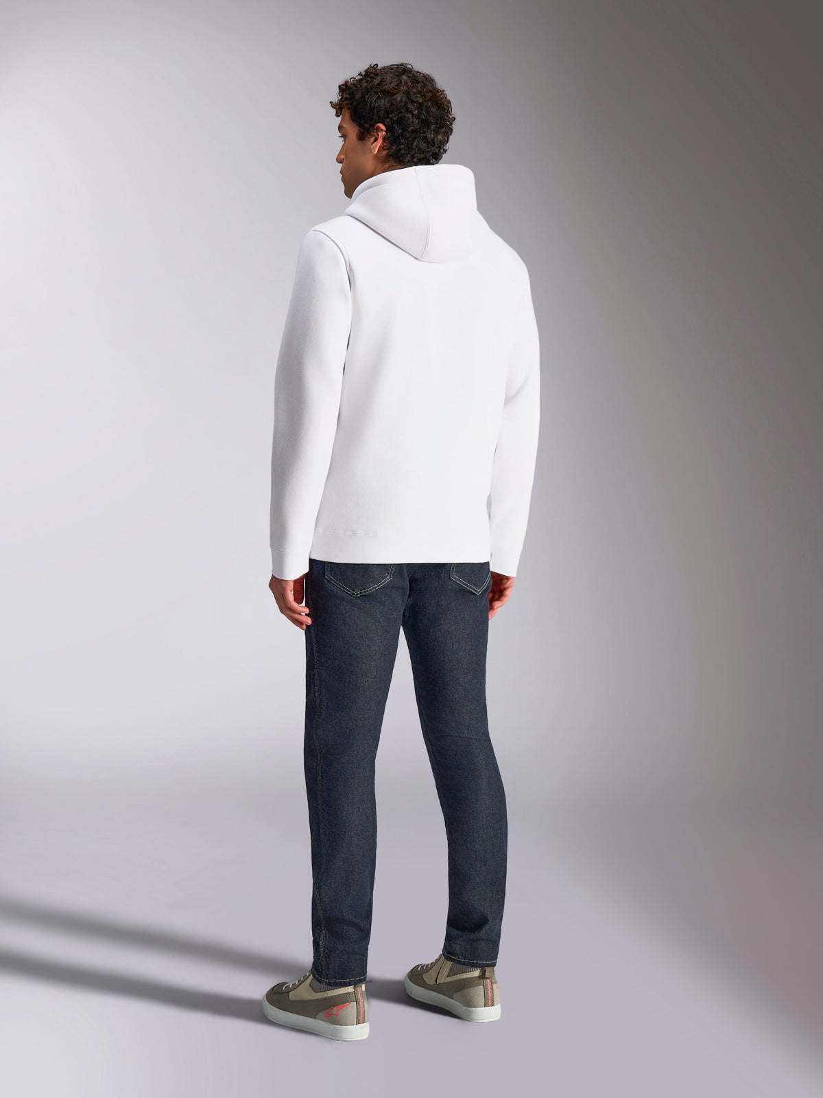 Sudadera Alpinestars Blaze V3, vista posterior de un modelo masculino que lleva una sudadera blanca con capucha, ropa deportiva casual combinada con vaqueros azul oscuro y zapatillas grises, diseño moderno inspirado en el automovilismo.