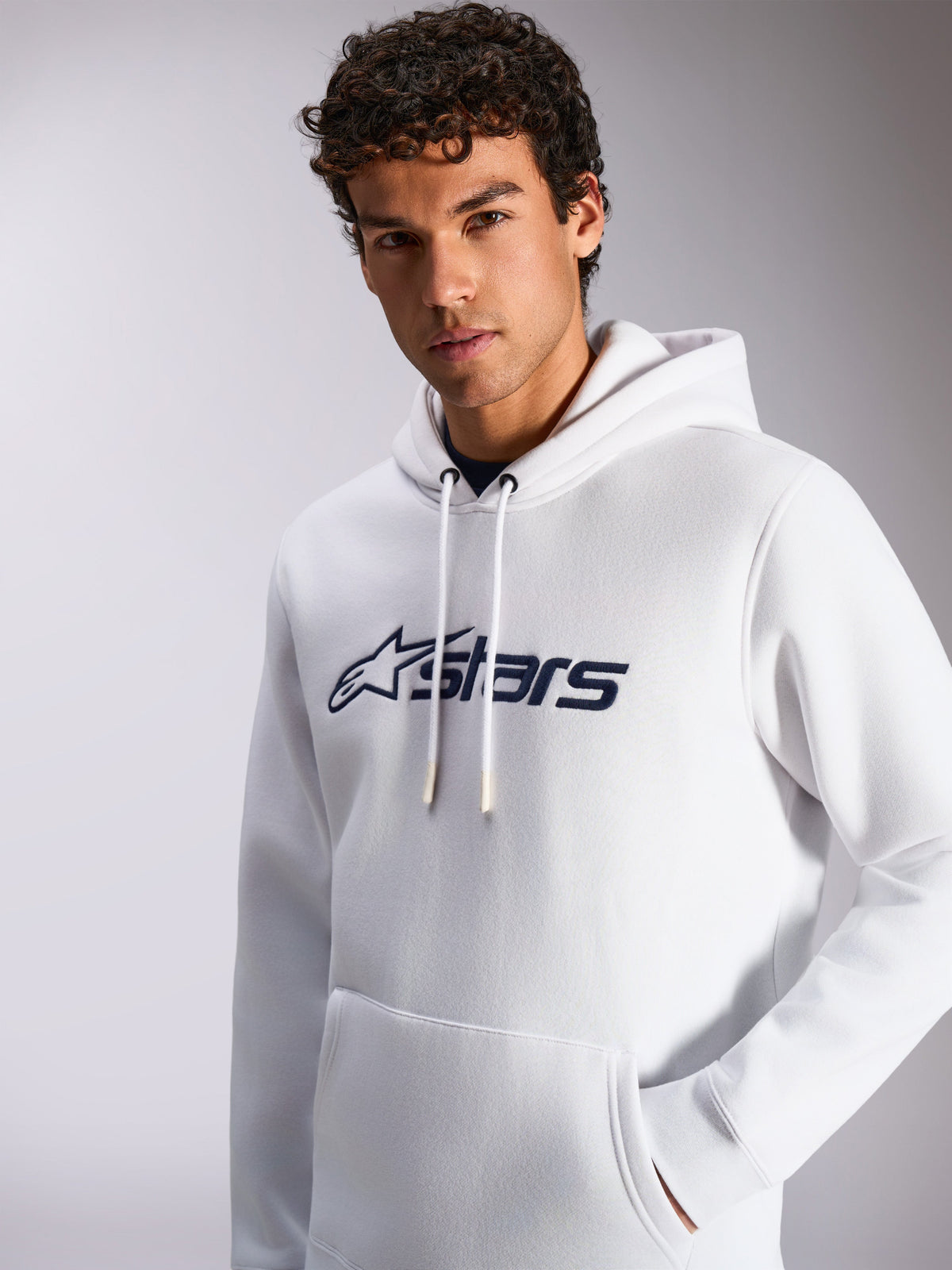 Sudadera Alpinestars Blaze V3, blanca y azul marino, usada por modelo masculino, ropa deportiva casual, logotipo frontal de Alpinestars en azul marino, capucha con cordón, bolsillo tipo canguro, diseño de estilo de vida moderno