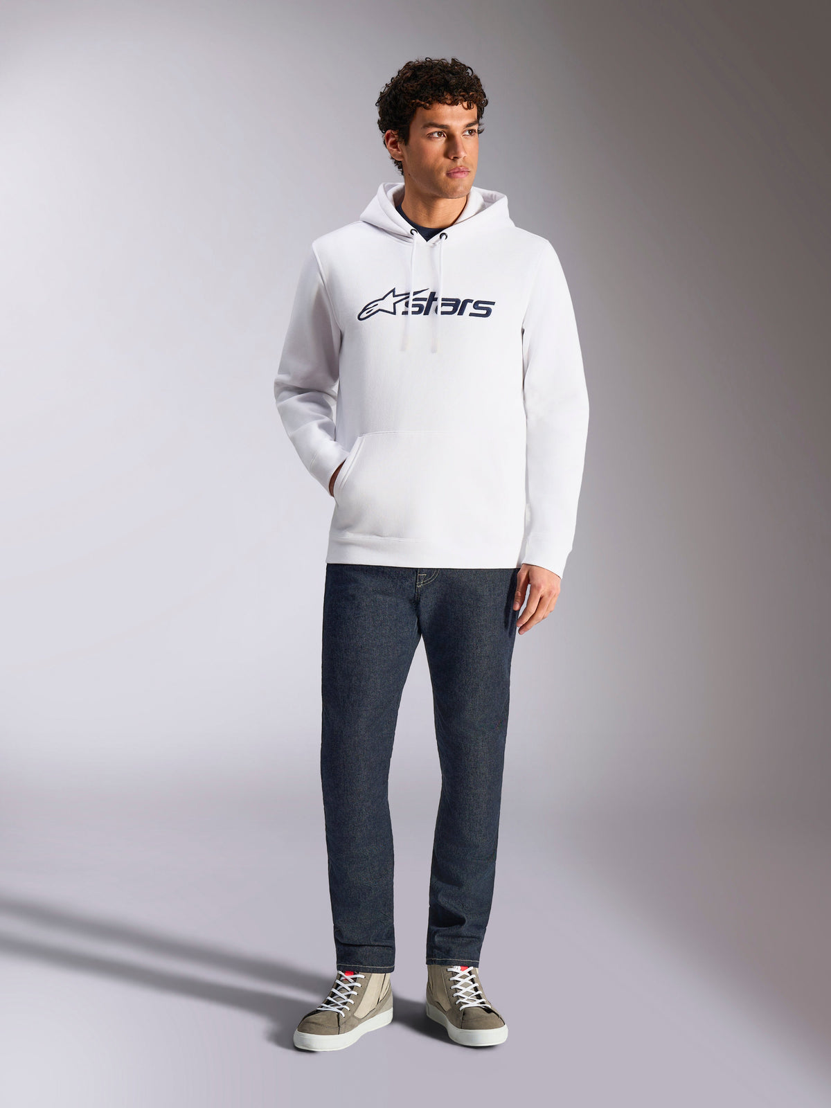 Sudadera con capucha Alpinestars Blaze V3, sudadera urbana casual blanca con el logotipo de Alpinestars en azul marino en el pecho, lucida por un modelo con vaqueros azul oscuro y zapatillas de color canela, con capucha con cordón y bolsillo de canguro frontal