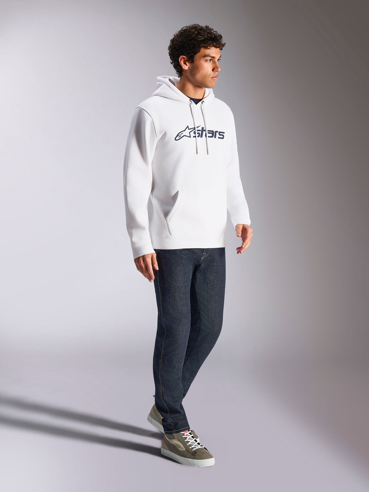 Sudadera con capucha Alpinestars Blaze V3, blanco y azul marino, estilo casual streetwear usado por un modelo con vaqueros azul oscuro y zapatillas, cuenta con capucha con cordón y un llamativo logotipo estampado en la parte delantera, diseño moderno inspirado en el automovilismo