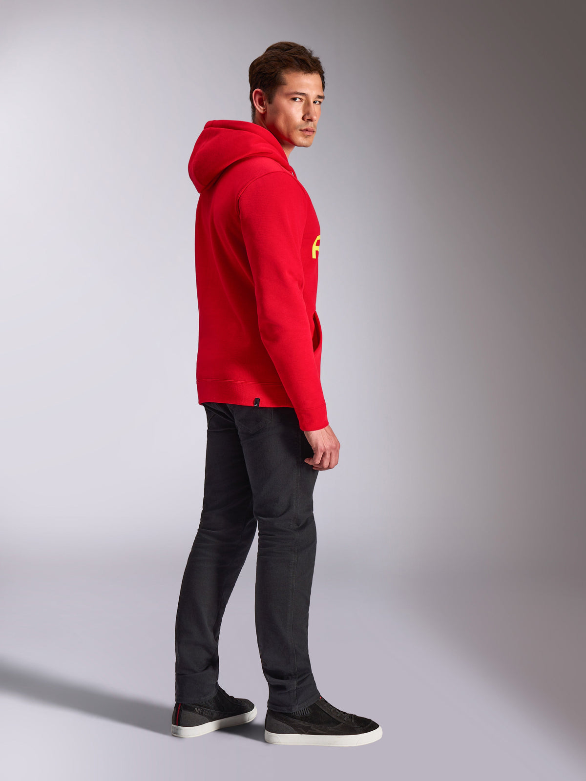 Sudadera con capucha Alpinestars Linear Wordmark V3, roja, ropa casual, modelo visto de perfil y desde atrás con una sudadera roja con logotipos amarillos, vaqueros gris oscuro y zapatillas negras, toma de estilo de vida en estudio