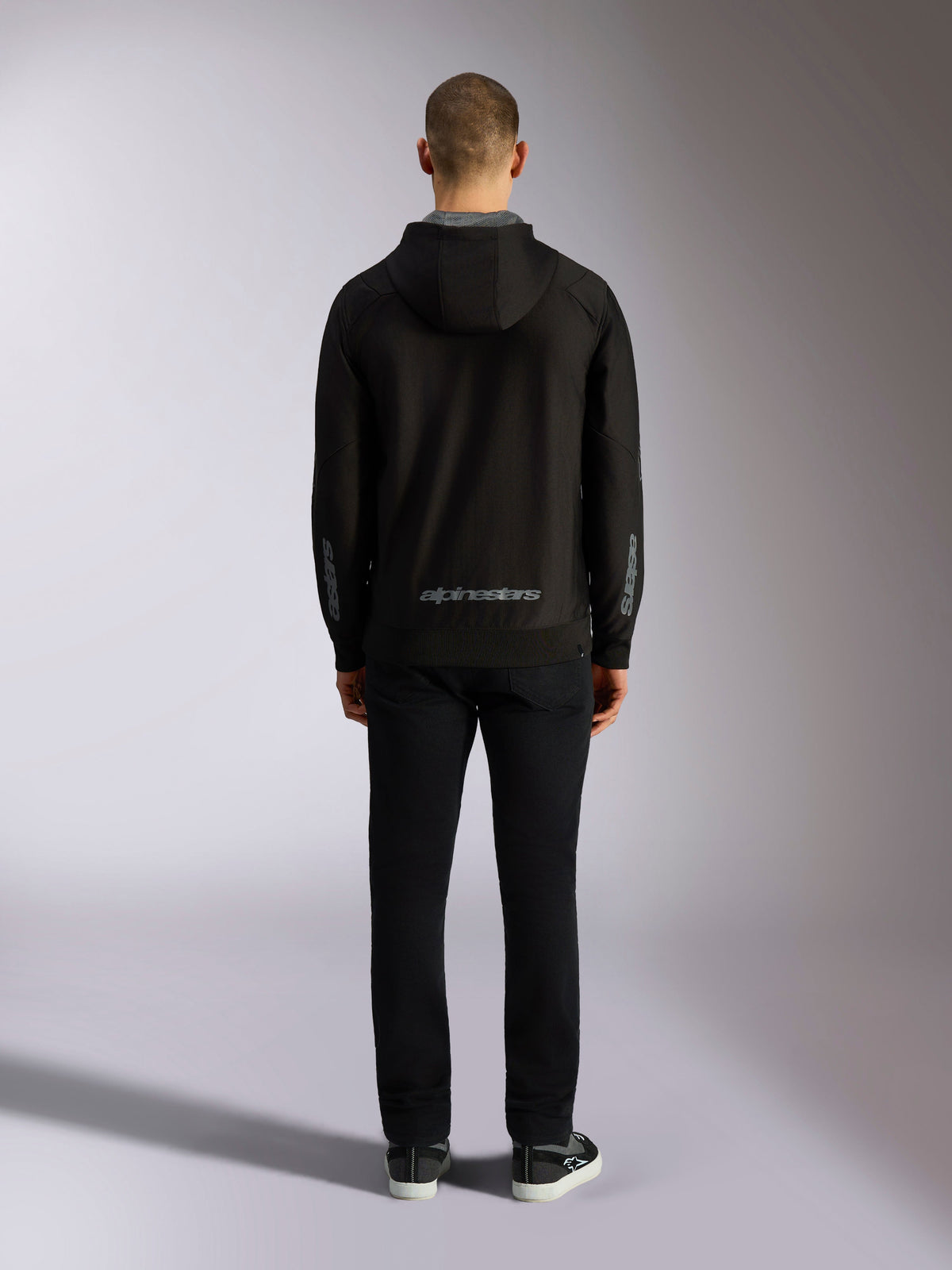 Vista trasera de la sudadera Alpinestars Sessions V3 Hoodie en color Black Grey, con la marca Alpinestars en gris en la zona inferior de la espalda y logotipos Astars en los antebrazos, vestida por un modelo con pantalones negros, estilo de ropa urbana casual inspirado en el motorsport
