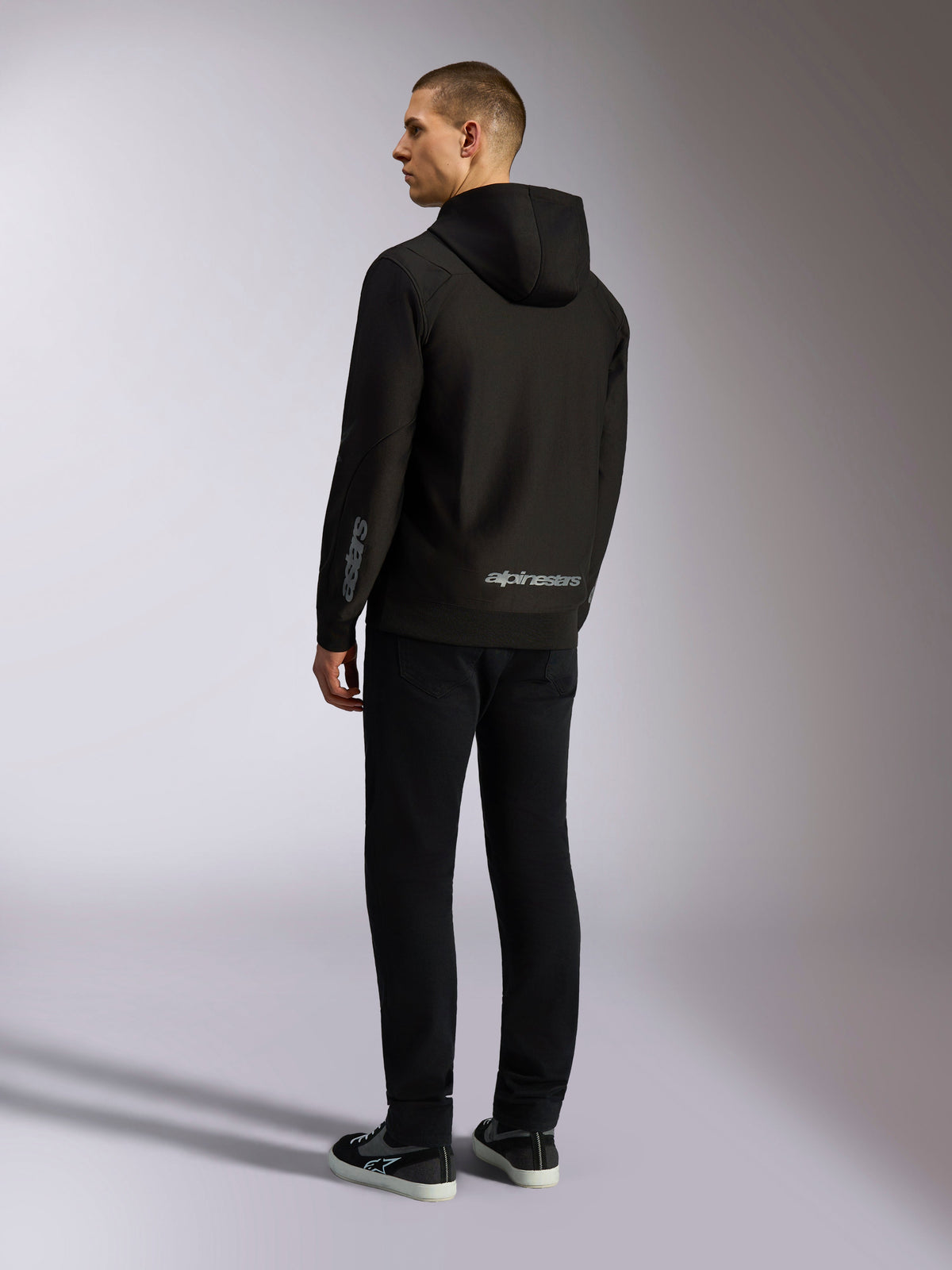 Sudadera Alpinestars Sessions V3, ropa urbana casual negra, vista trasera en un modelo, con la marca denominativa Alpinestars en gris en la parte inferior de la espalda y el logotipo Astars en la manga, diseño con capucha y cremallera frontal con estilo moderno inspirado en el automovilismo