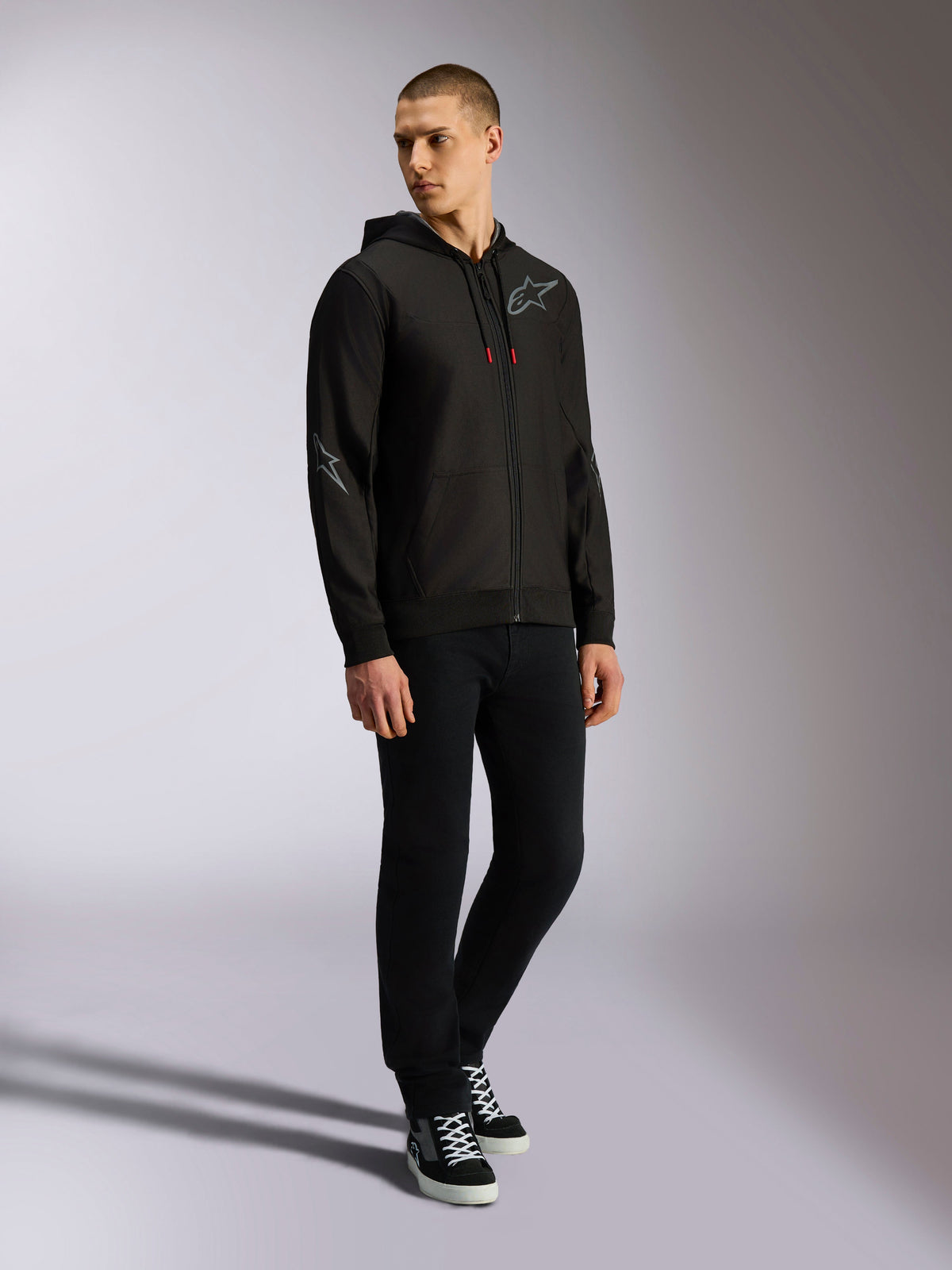 Sudadera Alpinestars Sessions V3 Hoodie, forro polar de poliéster con cremallera frontal, color negro y gris, logotipos grises en el pecho y las mangas, capucha con cordones de puntas rojas, vista de cuerpo completo de un modelo con ropa urbana de estilo motorsport
