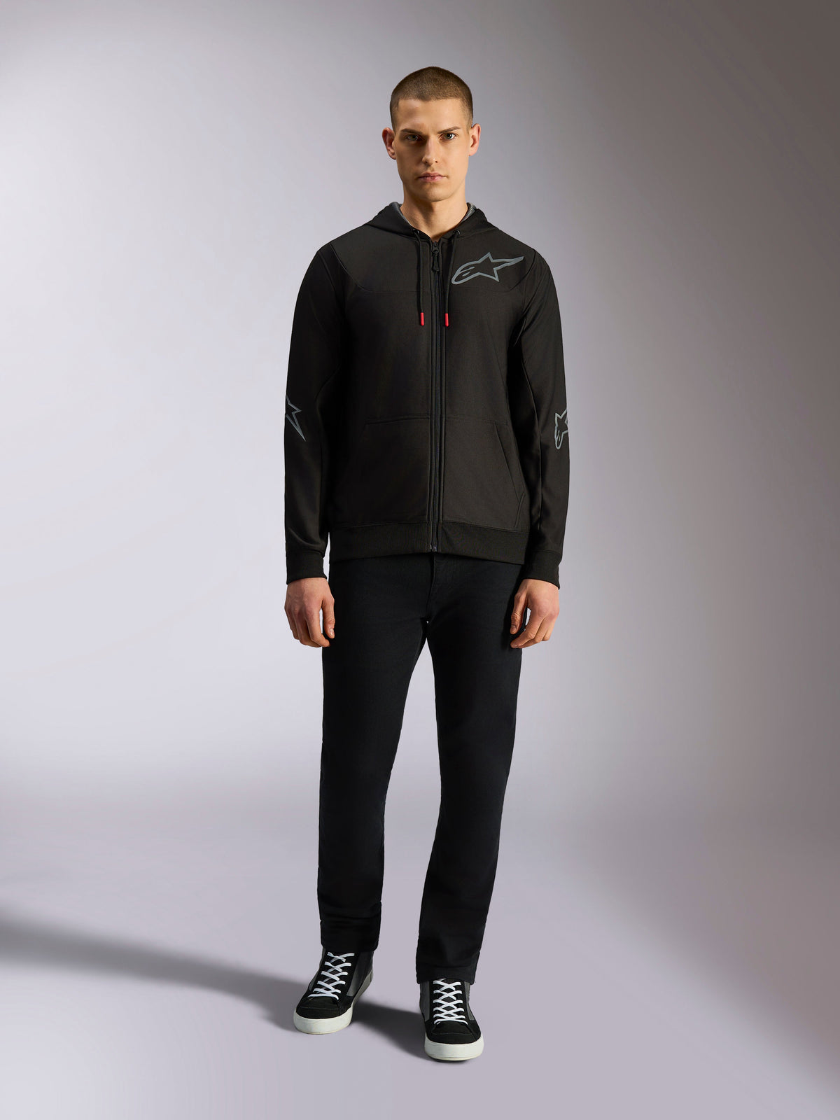 Alpinestars Sessions V3 Hoodie, sudadera negra con cremallera completa y logotipo gris de Astars en el pecho, gráficos en codos y antebrazos, detalles de cordones rojos, usada por modelo masculino con pantalones negros y zapatillas, ropa casual de motorsport