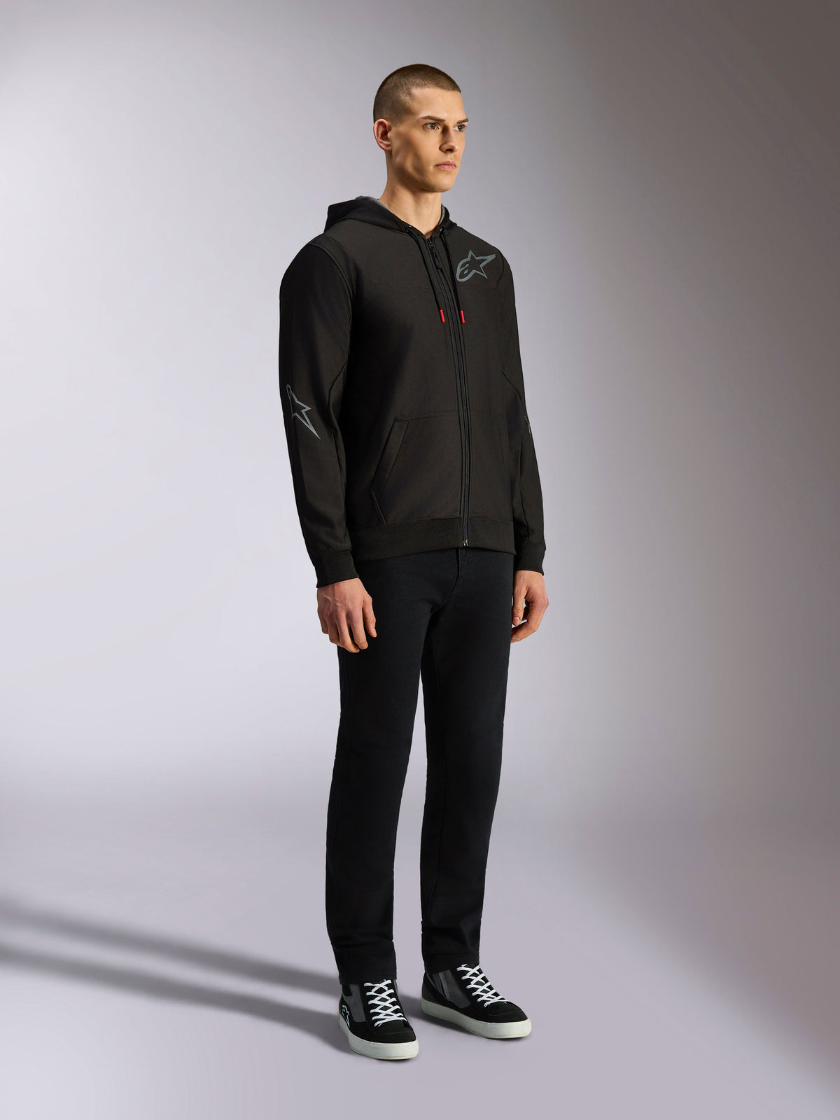 Sudadera Alpinestars Sessions V3, ropa urbana casual de automovilismo, sudadera negra con cremallera y logotipo de estrella gris en el pecho y la manga, cordones con puntas rojas, puños y dobladillo de canalé, lucida por un modelo con pantalones negros y zapatillas