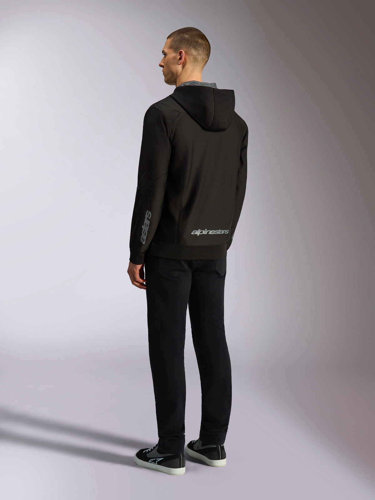 Sudadera con capucha Alpinestars Sessions V3, ropa casual de estilo motorsport, negra con logotipos grises en la parte inferior de la espalda y el antebrazo, forro polar de poliéster con cremallera frontal y capucha forrada de malla, vista trasera en modelo, diseño deportivo moderno