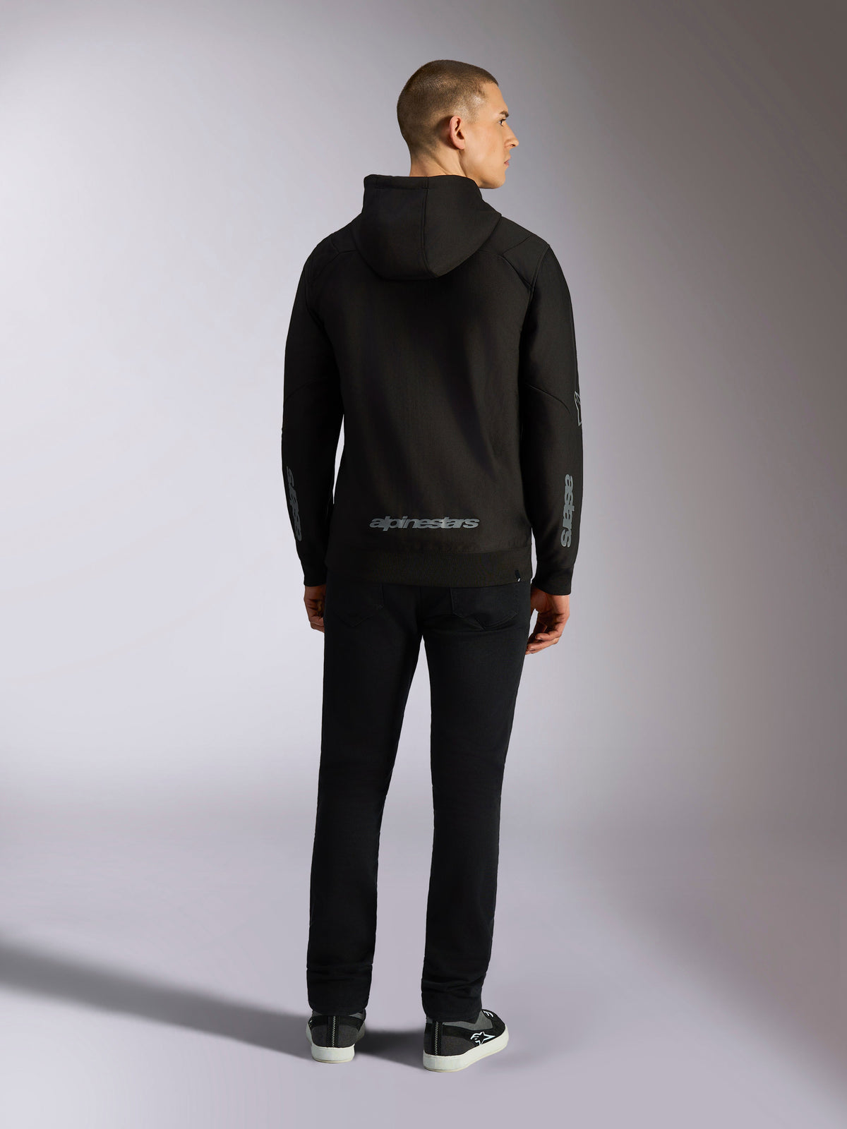 Alpinestars Sessions V3 Hoodie, streetwear casual, negro con logotipo Alpinestars gris en la parte inferior de la espalda y marca 'astars' en las mangas, vista posterior de modelo masculino con sudadera y pantalones negros, diseño moderno inspirado en el automovilismo