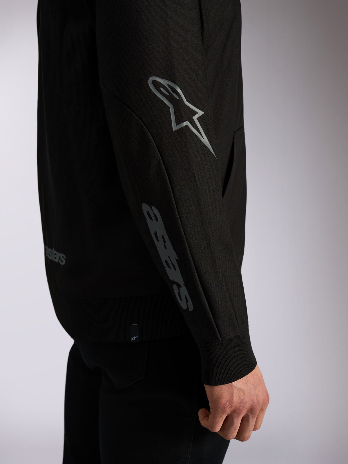 Sudadera con capucha Alpinestars Sessions V3, ropa urbana casual negra, primer plano de la manga con el logo Astars gris en el codo y la marca denominativa en el antebrazo, puños de canalé, diseño inspirado en el automovilismo