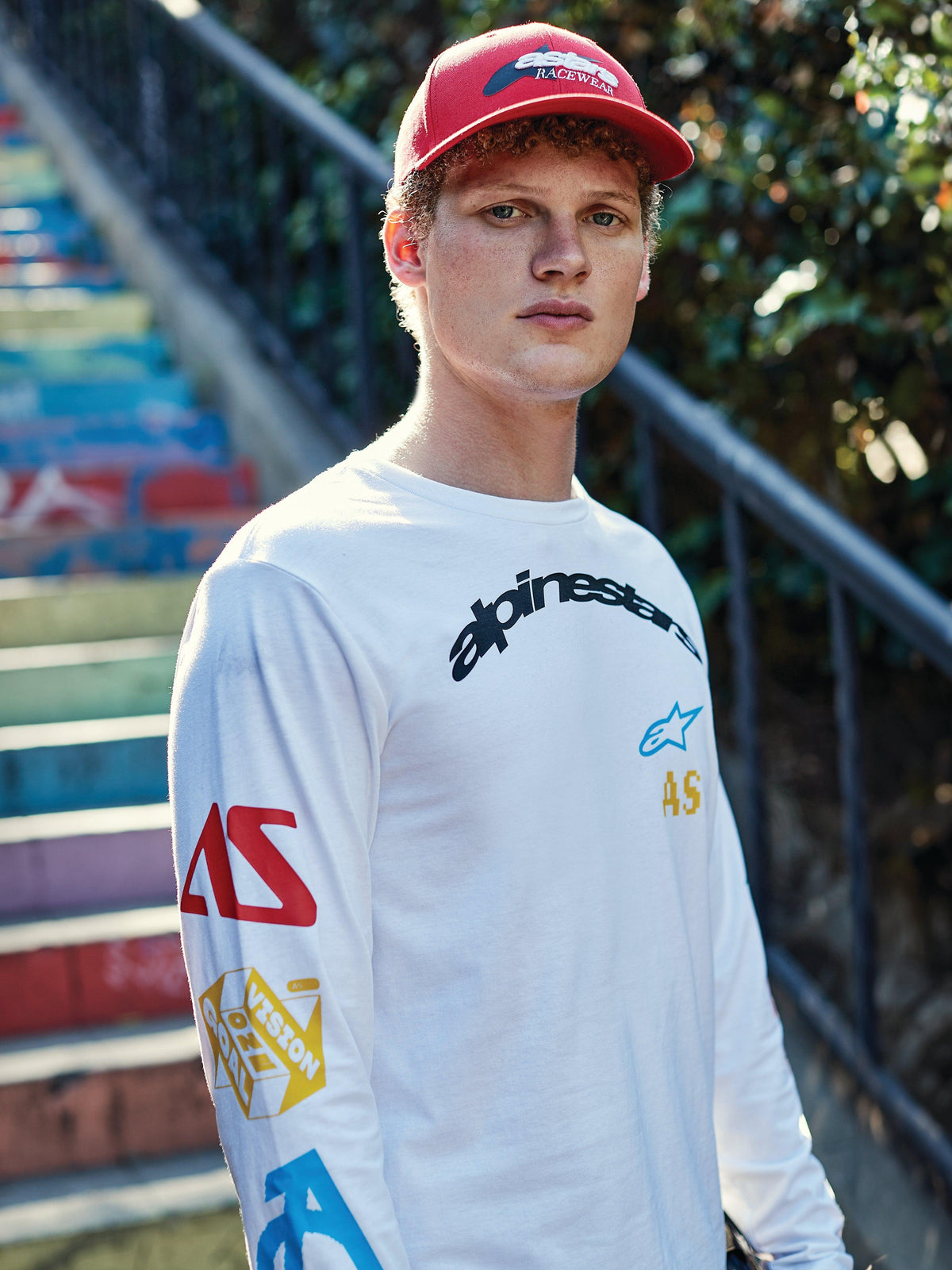 Alpinestars Brundo CSF Long Sleeve Tee, camiseta de manga larga blanca con logotipo negro en el pecho y gráficos multicolores en las mangas, vestida por un hombre con gorra roja, estilo casual motorsport streetwear