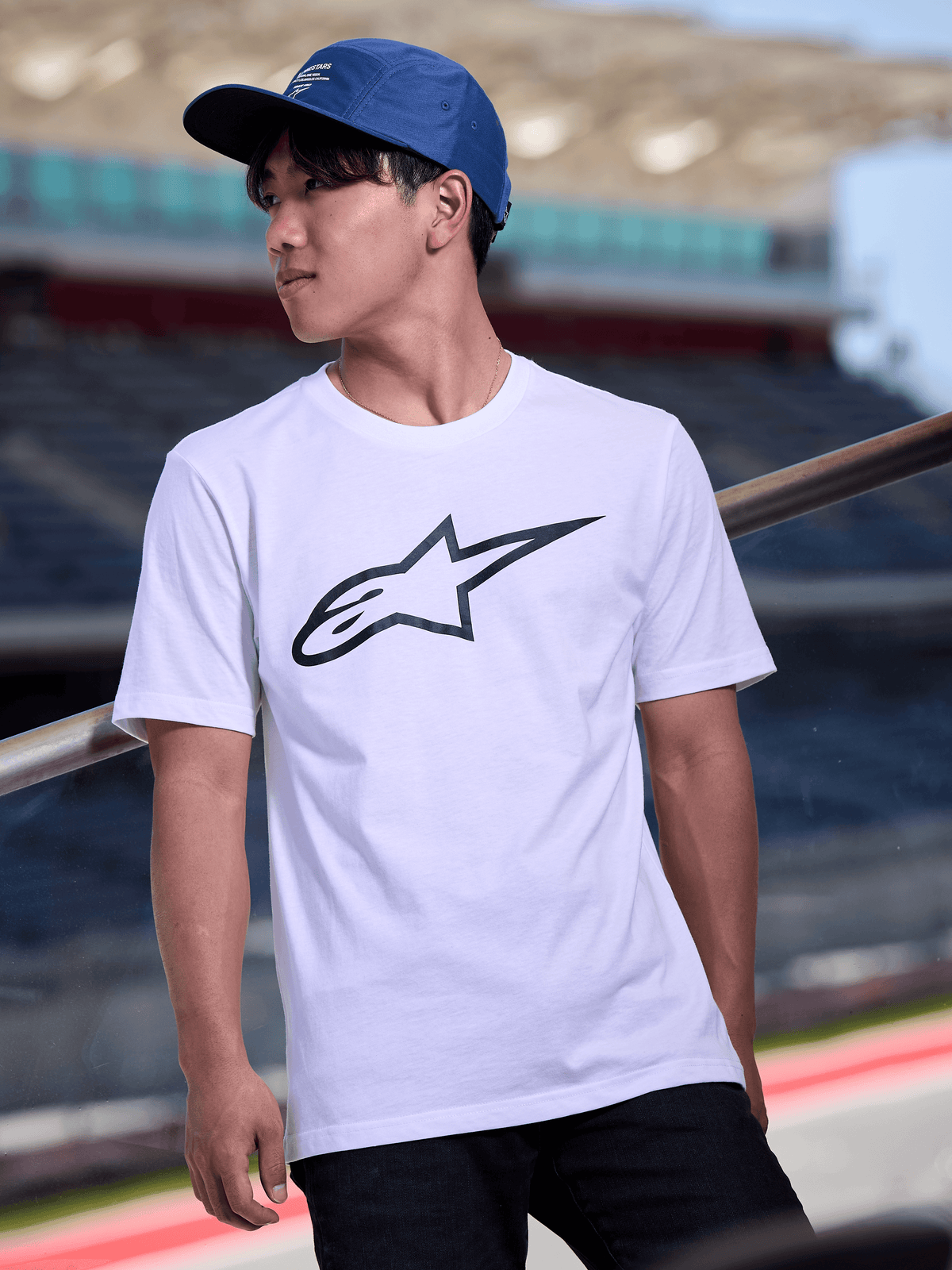 Hombre con la gorra Peasy de Alpinestars en color azul, gorra jockey de 5 paneles con visera plana y estampado gráfico del logotipo en blanco, combinada con una camiseta blanca con el logo de Alpinestars, estilo streetwear casual inspirado en el motor