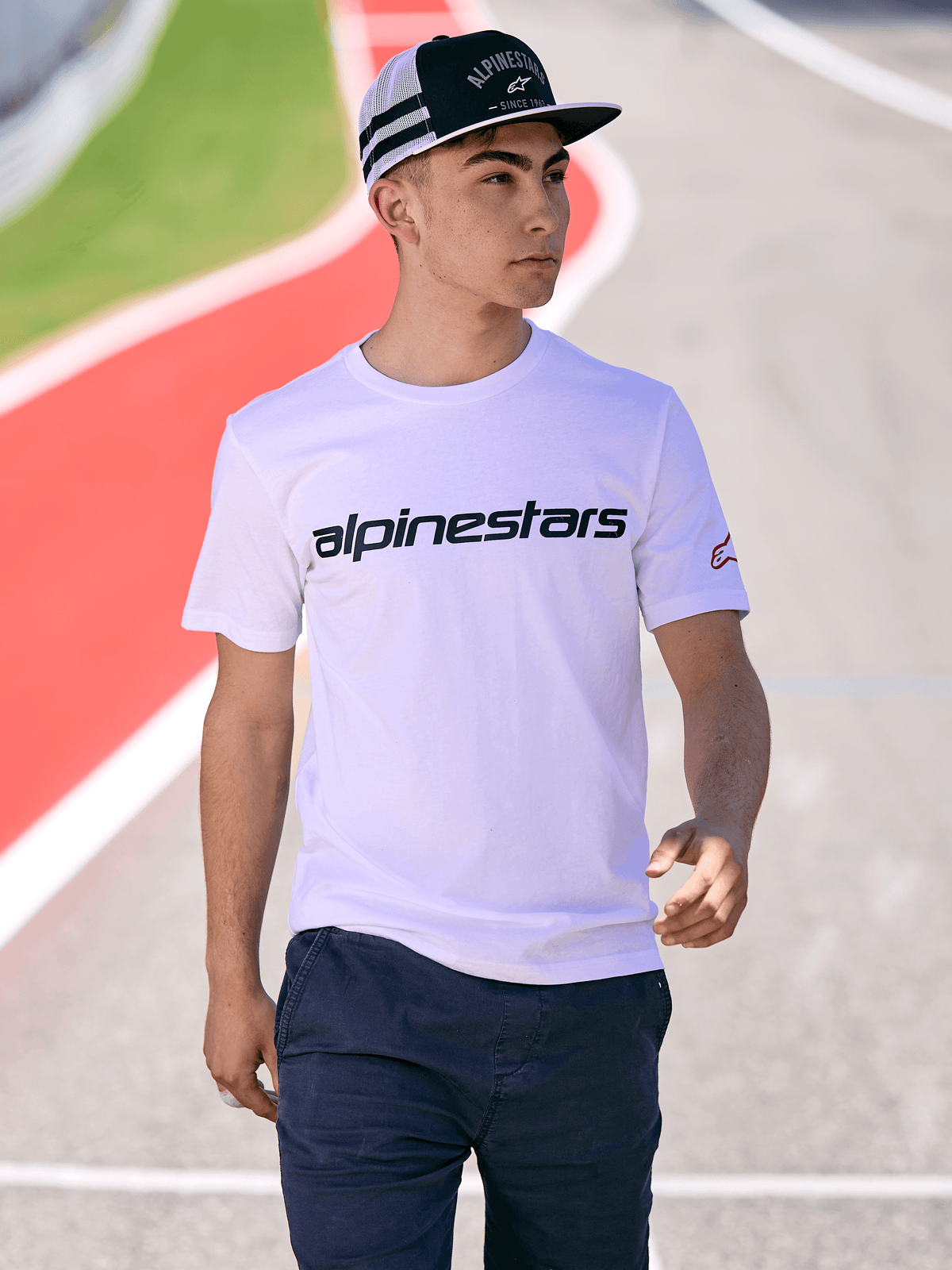 Hombre con gorra Trucker Backline de Alpinestars en blanco y negro, combinada con una camiseta blanca con el logotipo de Alpinestars y pantalones azul oscuro, estilo casual de motorsport en una pista de carreras