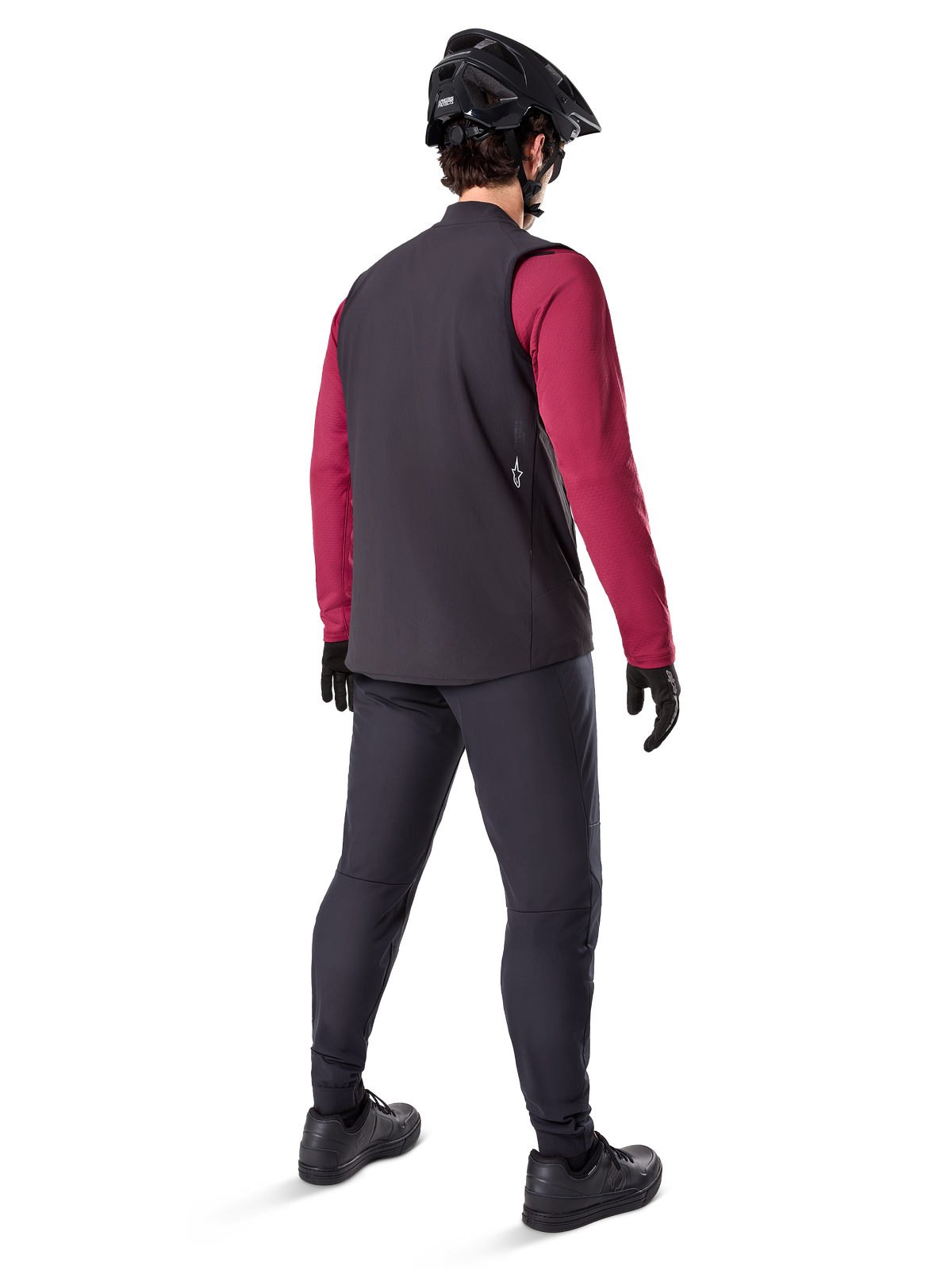 Chaleco Térmico Alpinestars A-Dura, chaleco de ciclismo negro, visto desde atrás en un modelo con un maillot granate de manga larga y pantalones de ciclismo negros, combinado con un casco de bicicleta de montaña y guantes negros, equipo técnico para rodar en climas fríos.