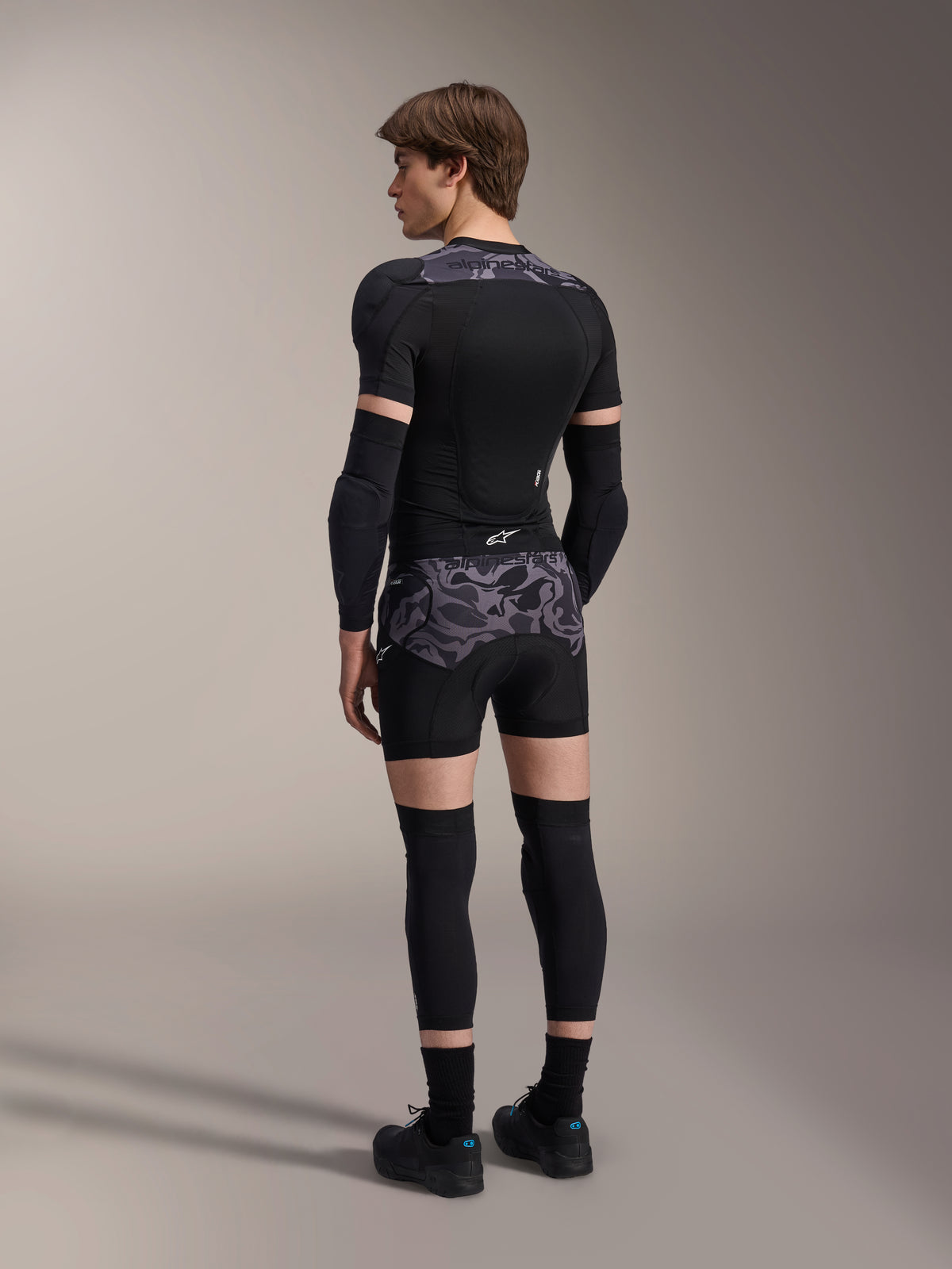 Modelo con boxers de protección Alpinestars A-Motion Plasma en Black Camo y chaqueta de protección A-Motion Plasma a juego, vista trasera que muestra el patrón de camuflaje gris, coderas, rodilleras y zapatillas de ciclismo, equipo completo de protección para bicicleta de montaña
