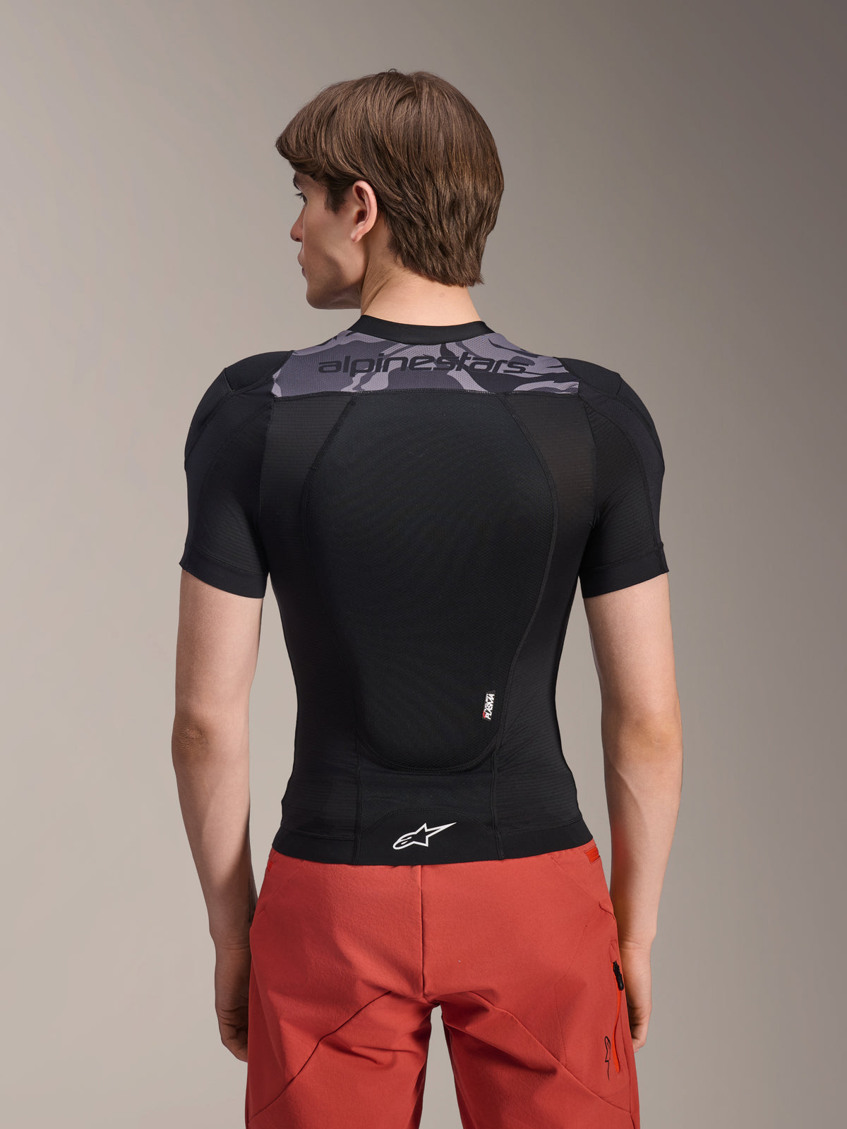 Chaqueta de protección Alpinestars A-Motion Plasma - Manga corta, negra con detalles de camuflaje gris en la parte superior de la espalda, vista trasera que muestra el protector de espalda integrado y logotipo de Astars blanco en el dobladillo, combinada con pantalones cortos rojos, equipo de protección técnica para bicicleta de montaña