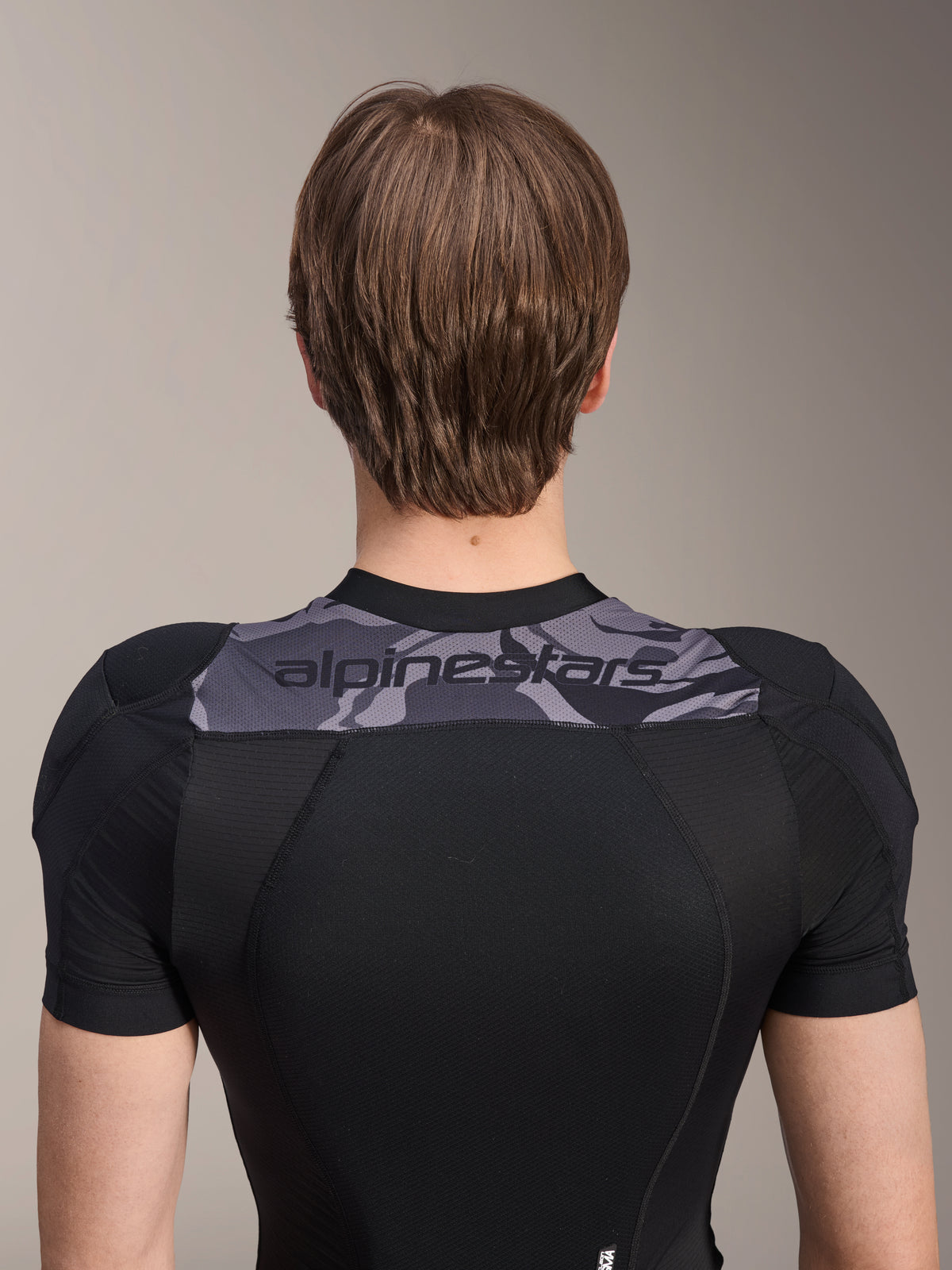 Chaqueta protectora Alpinestars A-Motion Plasma de manga corta, vista trasera de la parte superior del torso, negra con panel de camuflaje gris en los hombros con logotipo de Alpinestars, tejido de malla elástica transpirable para protección de MTB