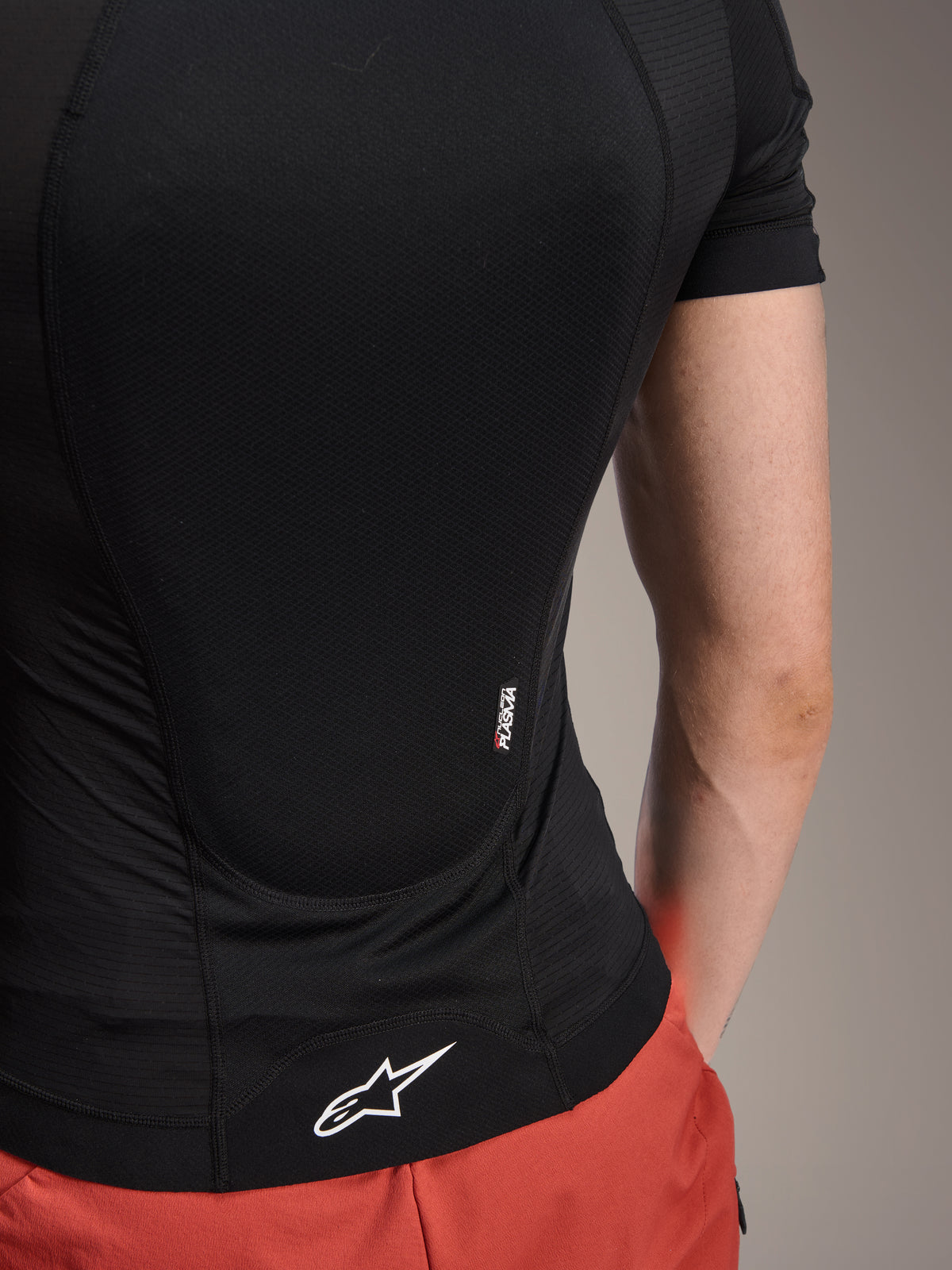 Alpinestars A-Motion Plasma Protection Jacket - Short Sleeve, camuflaje negro, detalle de la vista posterior con marca Nucleon PLASMA y logotipo de Alpinestars en el dobladillo, construcción de malla elástica y transpirable para protección en MTB