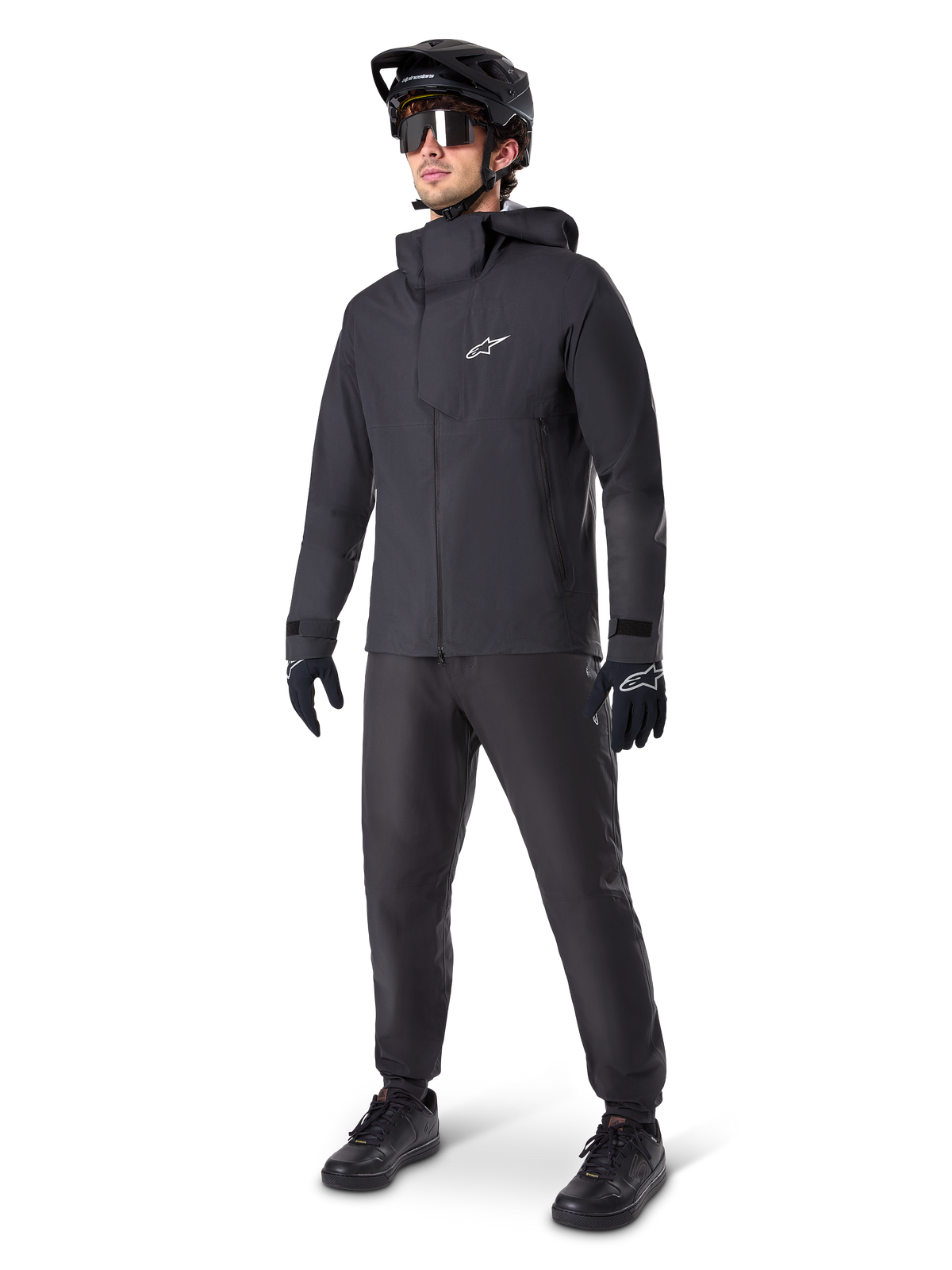 Male mountain biker wearing Alpinestars A-Dura Waterproof Pants in black, paired with a matching black hooded waterproof jacket, black MTB helmet with visor, dark cycling glasses, and black gloves, full professional gear set for all-weather trail riding
