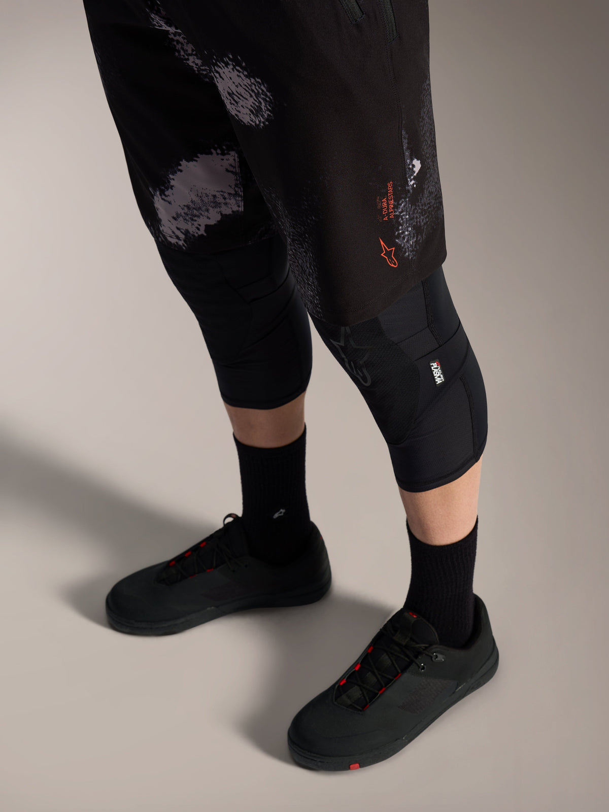 Pantalones cortos Alpinestars A-Dura Ride en Black Camo, equipo MTB con logo naranja, rodilleras y zapatillas.