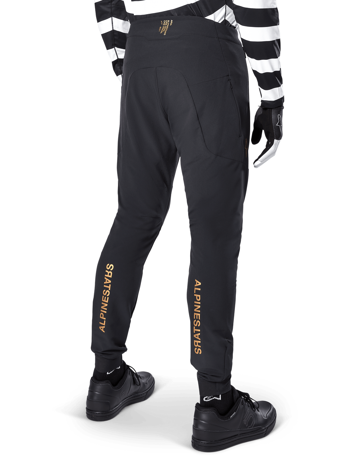 Pantalones Alpinestars A-Aria Elite, pantalones de MTB negros, vista trasera, con la marca Alpinestars en dorado vertical en la parte posterior de las piernas, usados con un jersey a rayas blancas y negras y calzado negro, ropa de ciclismo de alto rendimiento para mountain bike