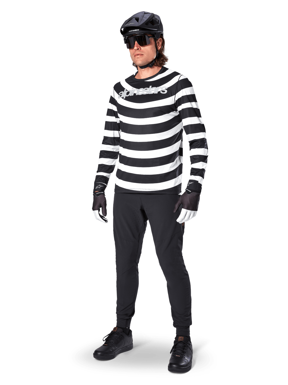 Pantalones Alpinestars A-Aria Elite, pantalones de MTB negros, usados por un ciclista con un jersey de manga larga a rayas blancas y negras, casco negro, gafas de sol y guantes blancos y negros, equipamiento de alto rendimiento para ciclismo todoterreno