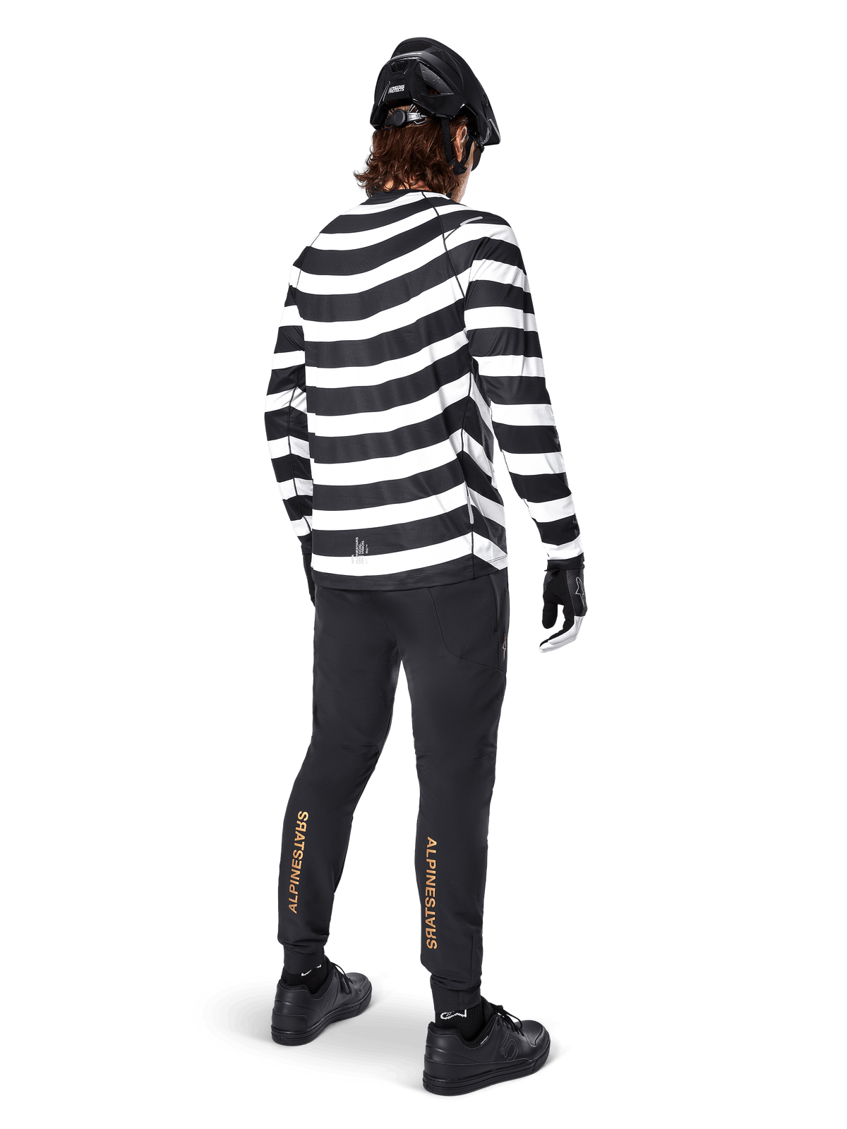 Pantalones Alpinestars A-Aria Elite, pantalones de MTB negros con un logotipo vertical dorado en las pantorrillas, usados por un ciclista con un jersey de manga larga a rayas blancas y negras, casco negro y zapatillas negras, vista posterior que muestra el ajuste de rendimiento y el diseño para senderos