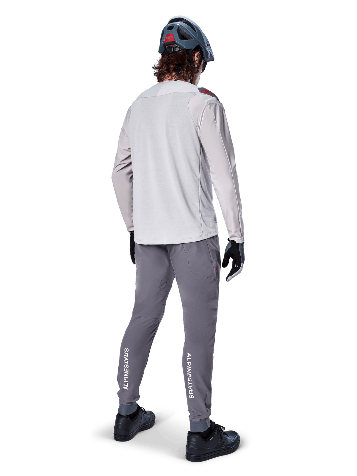 Pantalones Alpinestars A-Aria Elite, pantalones de ciclismo MTB, gris oscuro, vista trasera de un ciclista con la equipación completa, con logotipos blancos de Alpinestars en la parte inferior de las piernas, ajuste de rendimiento con perforaciones láser, combinados con un jersey de manga larga gris claro y casco de MTB gris