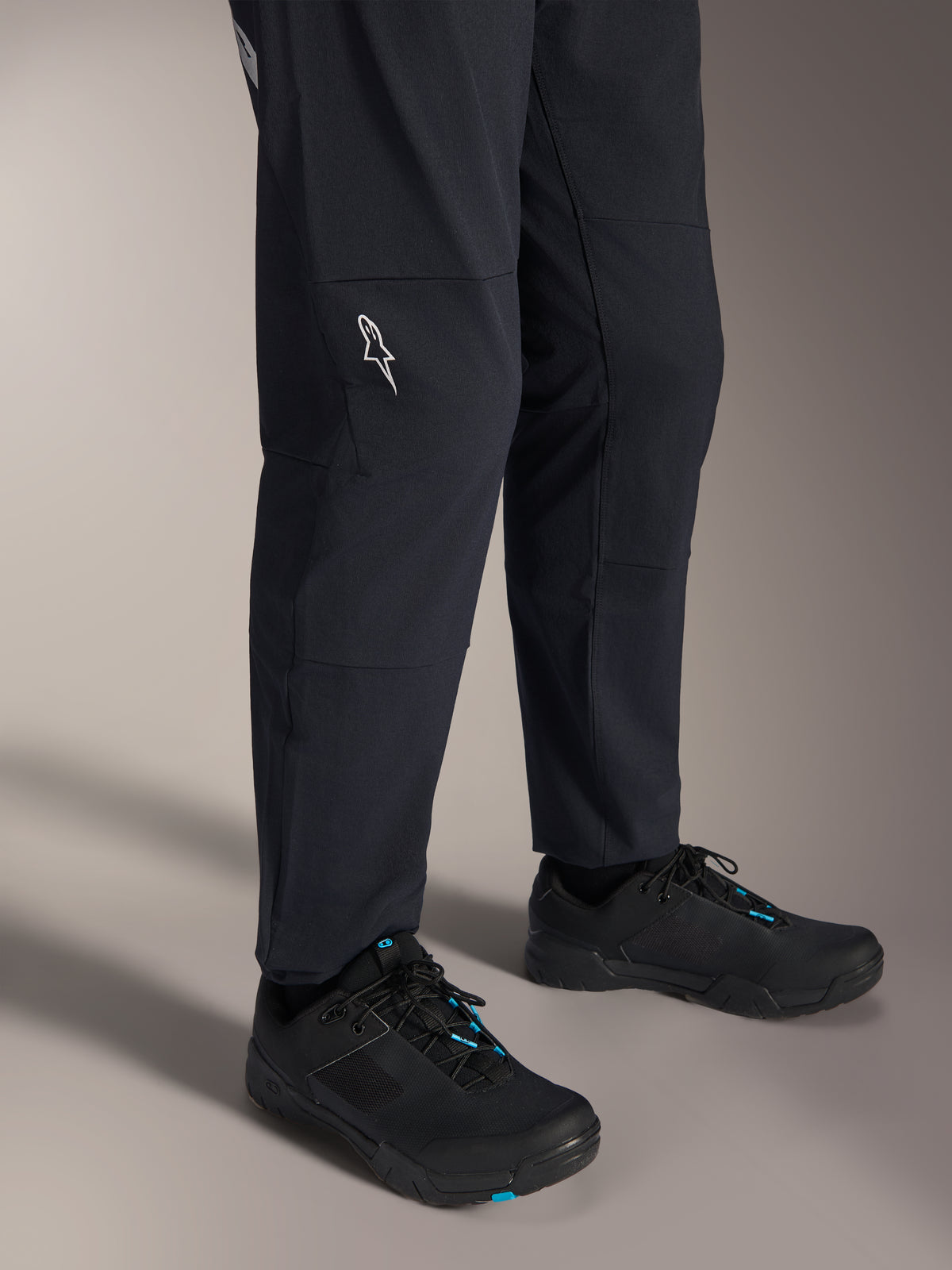 Pantalones Alpinestars A-Dura Elite, pantalones negros para bicicleta de montaña, vista de la parte inferior de la pierna con un logotipo blanco de Alpinestars en la rodilla, usados con zapatillas de ciclismo negras y azules, construcción duradera para MTB