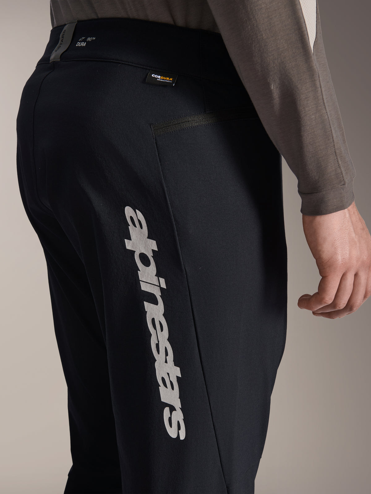 Pantalones Alpinestars A-Dura Elite, pantalones negros de ciclismo de montaña, vista lateral que muestra el gran logotipo vertical de Alpinestars en la pierna, etiqueta de tejido Cordura y bolsillo lateral con cremallera, ropa técnica de ciclismo para rutas off-road.