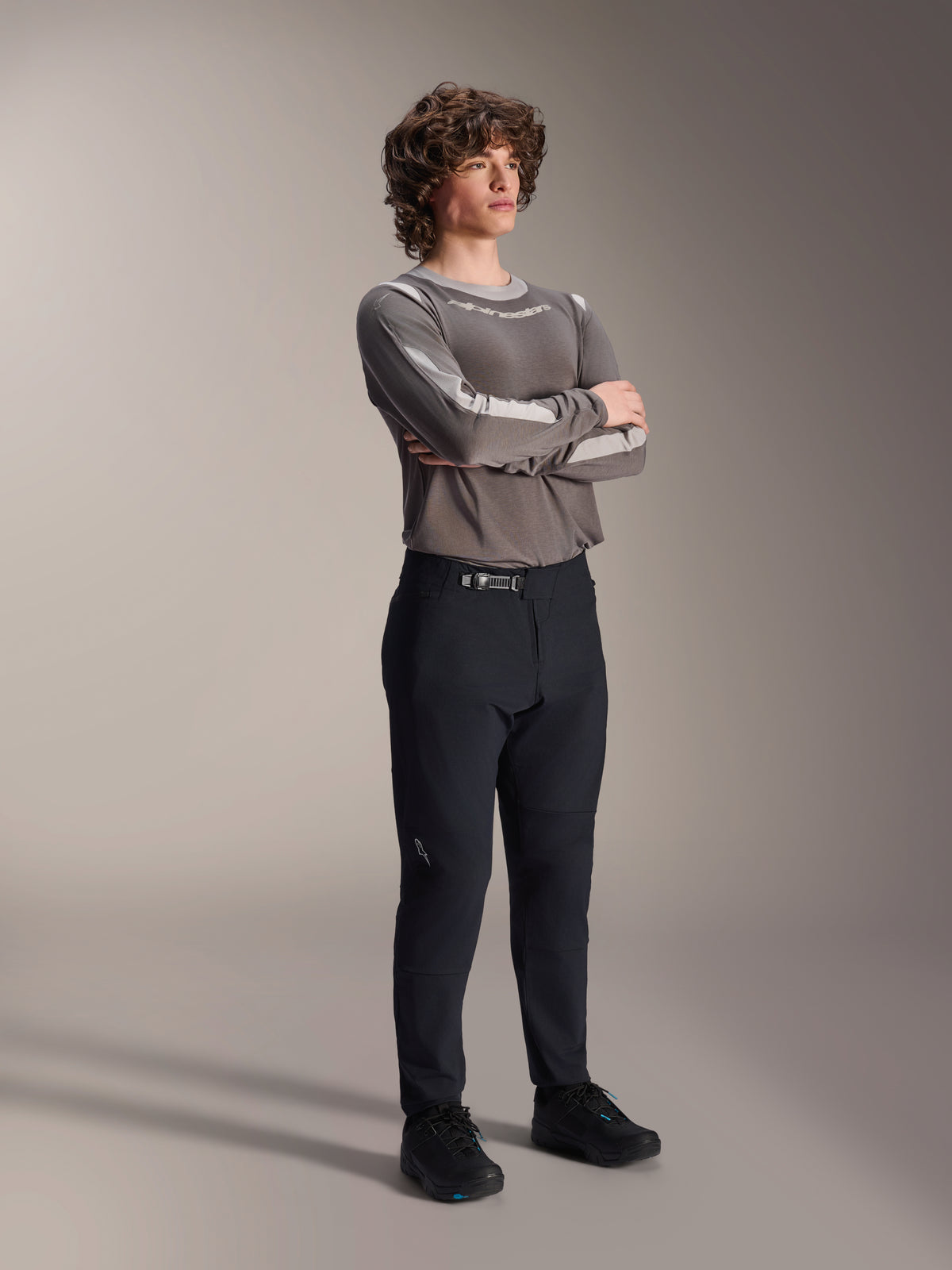 Modelo masculino con pantalones Alpinestars A-Dura Elite, pantalones negros de MTB, combinados con un jersey gris de manga larga y zapatillas de ciclismo negras, sistema de cierre de trinquete frontal, ropa profesional de bicicleta de montaña para trail y enduro
