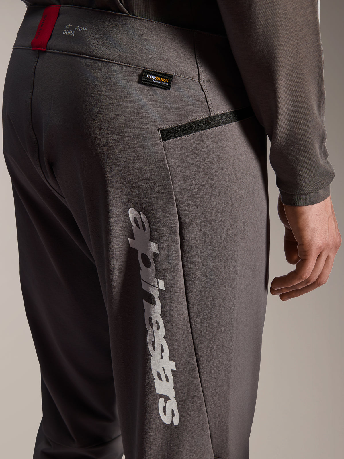 Pantalones Alpinestars A-Dura Elite, pantalones de MTB gris oscuro, vista de cerca de la cadera y el lateral del muslo, con un gran logotipo blanco vertical de Alpinestars, etiqueta de tejido Cordura, bolsillo con cremallera y detalle en rojo en la cintura