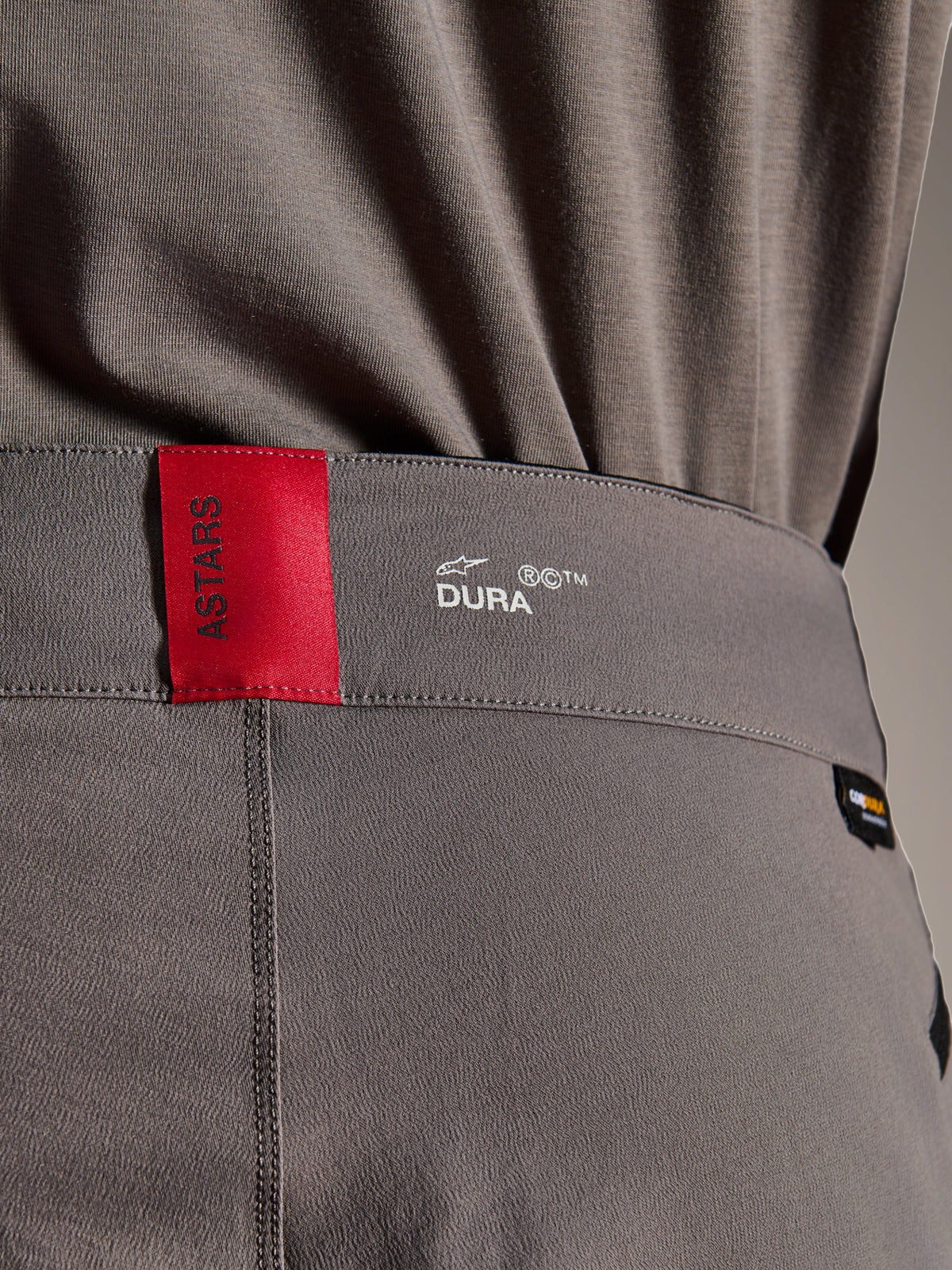 Alpinestars A-Dura Elite Pants, pantalones de MTB, gris oscuro, primer plano de la cintura que muestra una etiqueta ASTARS roja y el logo DURA blanco, con tejido Cordura texturizado y detalles de marca premium