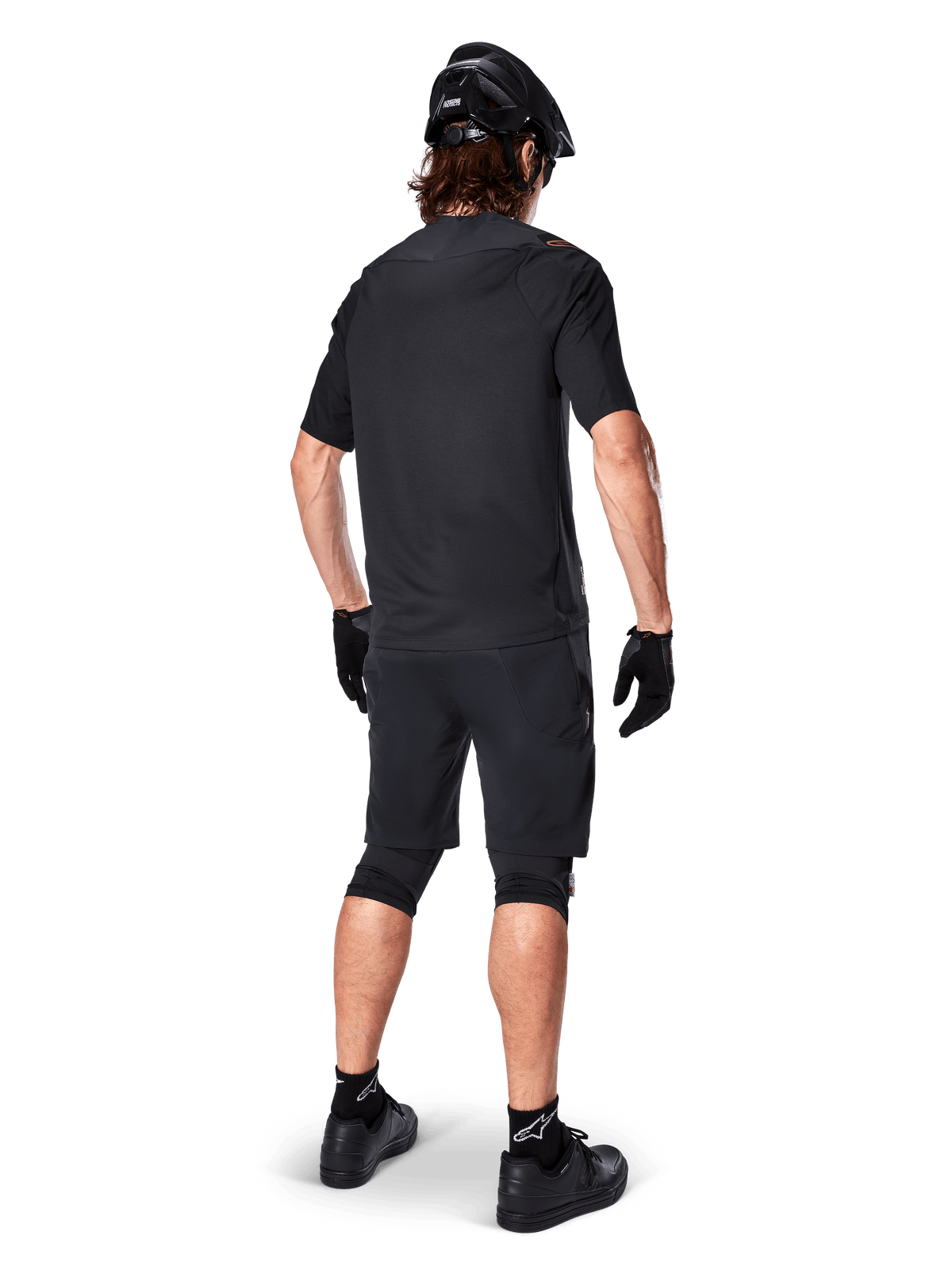 Rear view of a mountain biker wearing Alpinestars A-Aria Elite Shorts in Black, paired with a matching black short-sleeve jersey, black helmet, gloves, and MTB shoes, showing a complete performance outfit for off-road cycling.