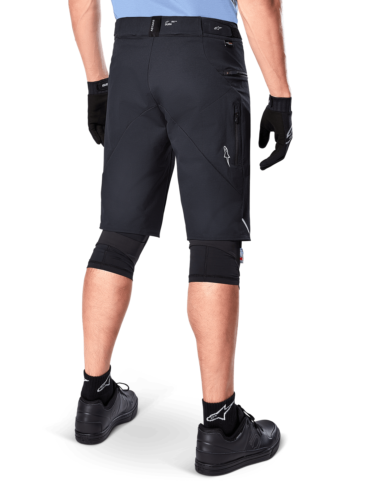 Shorts Alpinestars A-Dura Elite, negros, vista trasera de un ciclista con equipo de bicicleta de montaña que incluye rodilleras negras, guantes y calcetines con logos blancos, con bolsillos laterales con cremallera y detalles reflectantes en un tejido Cordura resistente.
