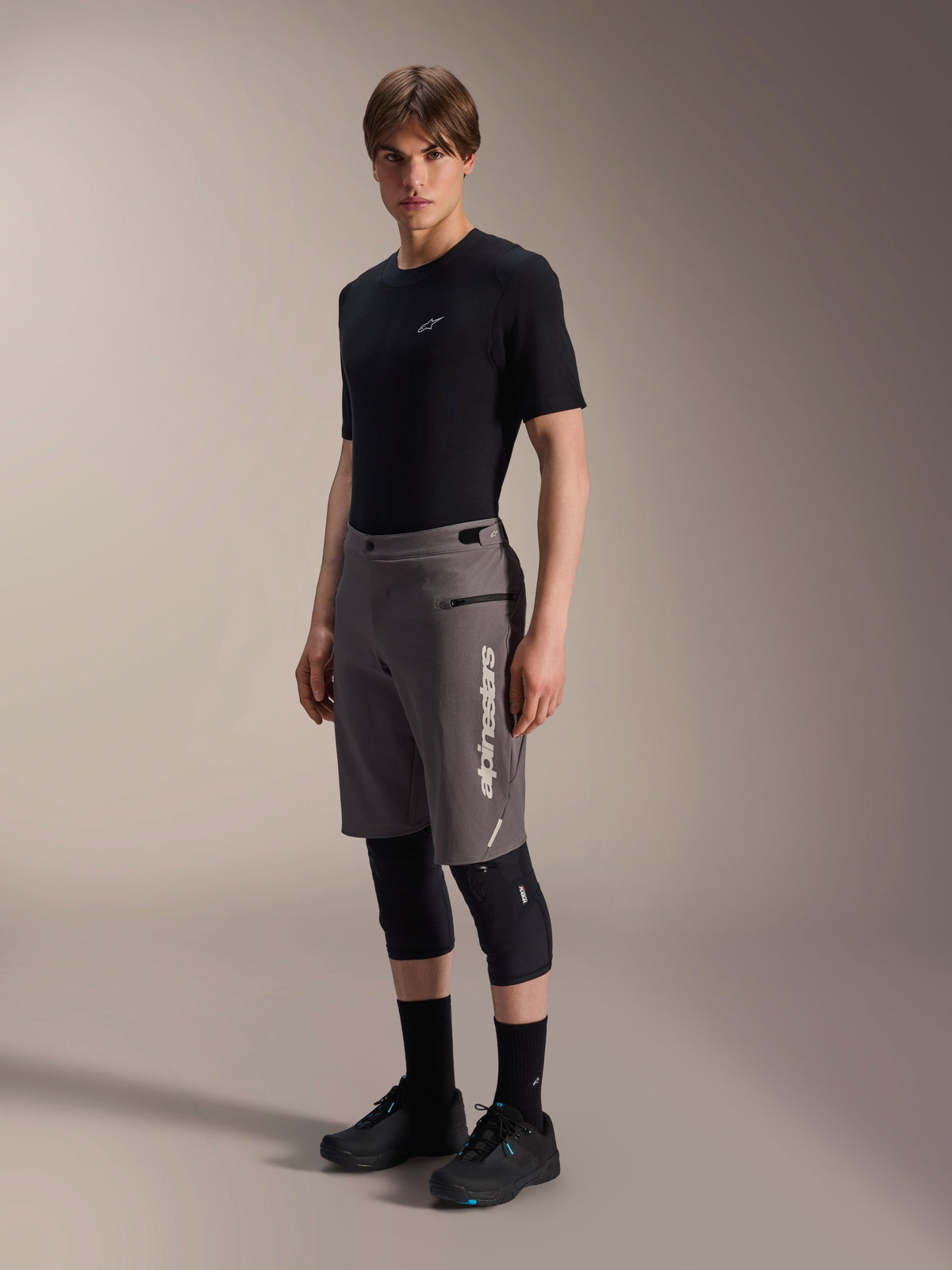 Alpinestars A-Dura Elite Shorts, Dark Gray mountain bike shorts, worn by a model with a black t-shirt and knee guards, featuring a vertical white Alpinestars logo on the left leg, technical Cordura fabric for off-road cycling.