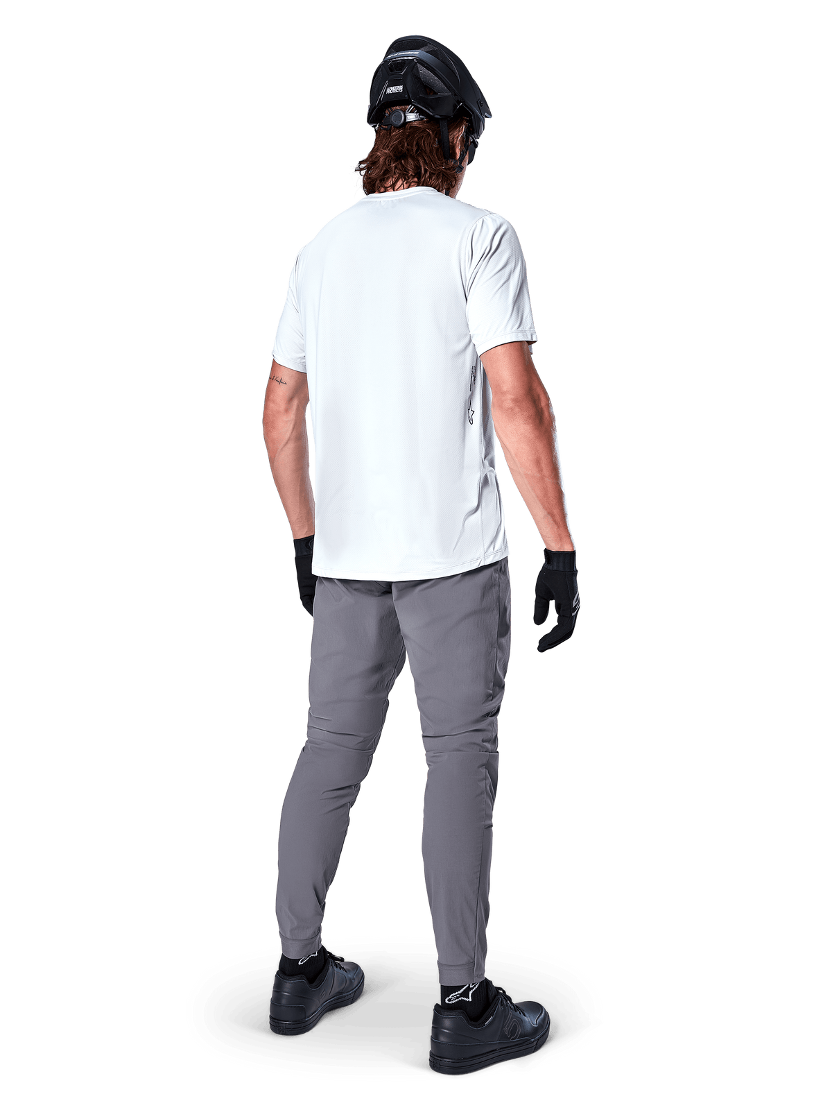 Pantalones Alpinestars A-Dura, pantalones de mountain bike gris oscuro vistos desde atrás, usados con una camiseta blanca de manga corta, casco de MTB negro, guantes y zapatillas negros, diseño de ajuste regular con puños elásticos, equipamiento de ciclismo duradero para todoterreno