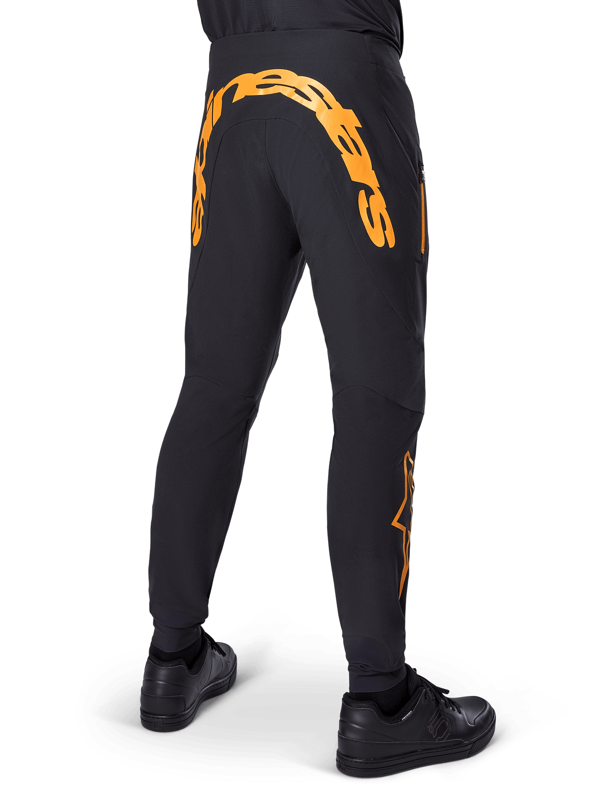 Alpinestars A-Supra Race Pants, pantalones de MTB negros, vista trasera con una gran marca denominativa Alpinestars en naranja en el canesú trasero, logotipo naranja en la parte inferior de la pierna, ajuste de alto rendimiento con bolsillo con cremallera, usados con zapatillas negras