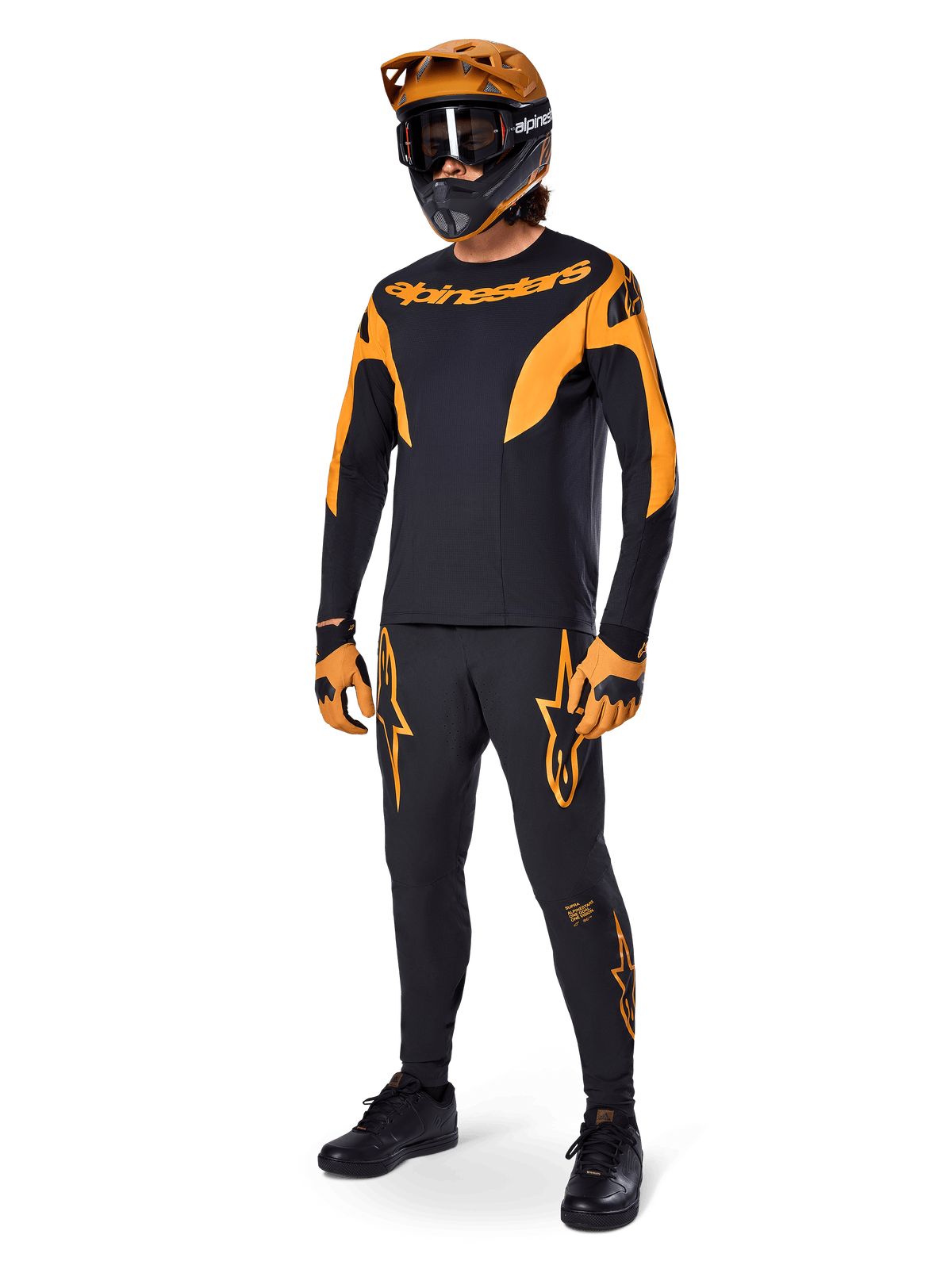 Ciclista de montaña con equipamiento completo de competición, pantalones Alpinestars A-Supra Race en negro con detalles naranjas, jersey de manga larga negro y naranja a juego, casco naranja con gafas negras, guantes naranjas y negros y calzado negro, diseño de kit de competición profesional de MTB