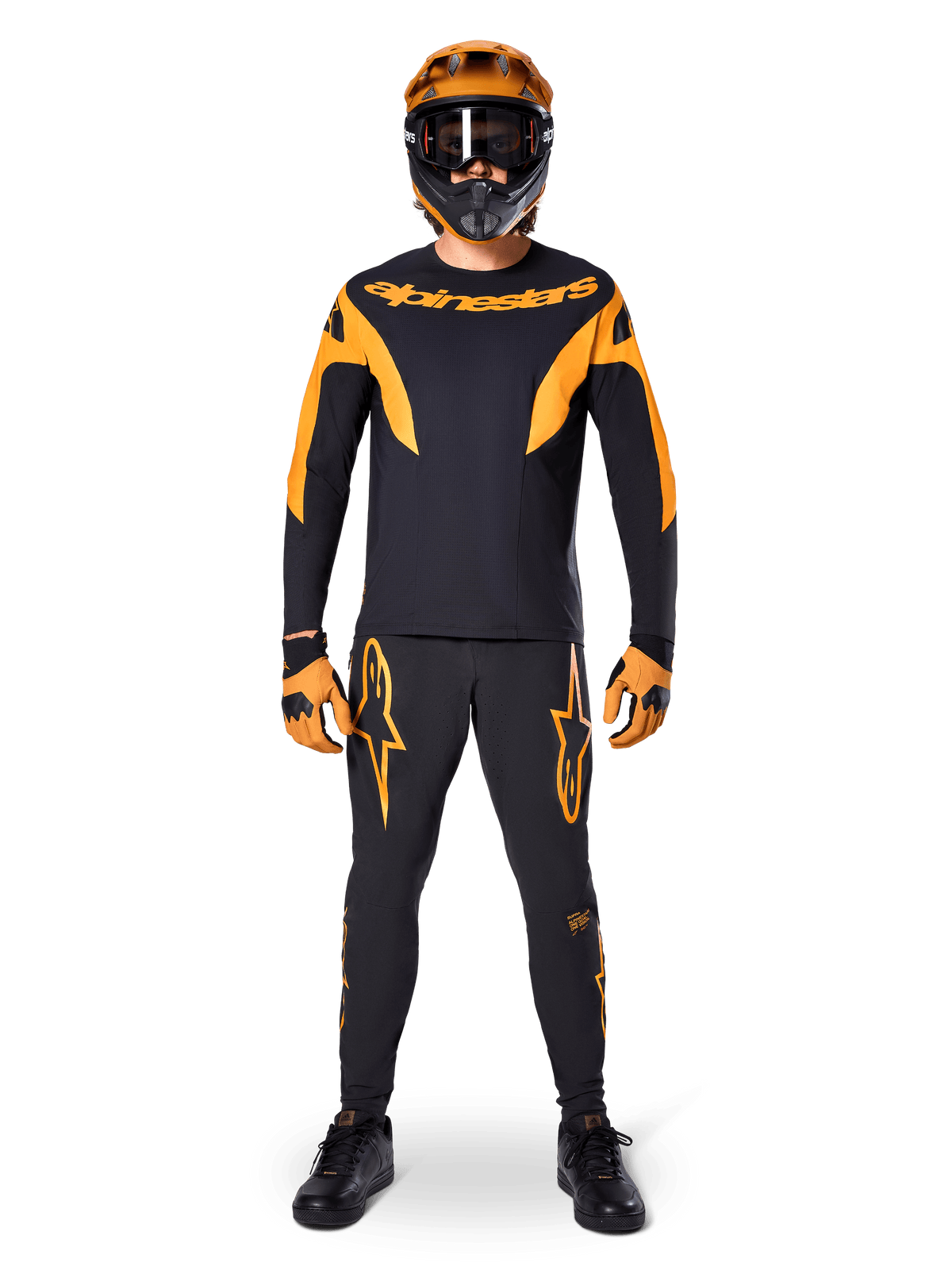 Pantalones Alpinestars A-Supra Race, pantalones de ciclismo de montaña, negros y naranjas, usados por un piloto con un equipamiento de carreras completo que incluye jersey negro y naranja, casco naranja y guantes a juego, con llamativos logotipos de estrella naranjas en las piernas