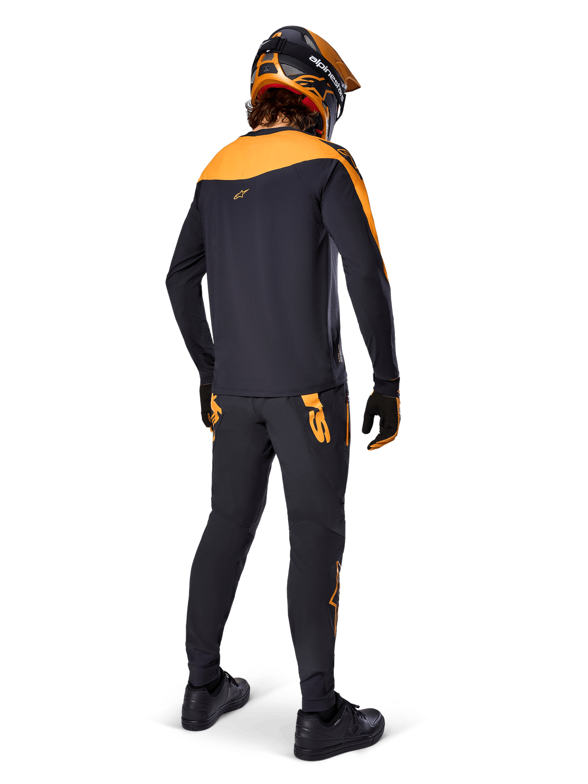 Pantalones Alpinestars A-Supra Race, pantalones de competición MTB, negros con logotipos naranjas, vistos desde atrás en un ciclista con jersey, casco y guantes negros y naranjas a juego, resaltando el logotipo inspirado en MX en el canesú trasero y el ajuste de rendimiento para senderos técnicos.