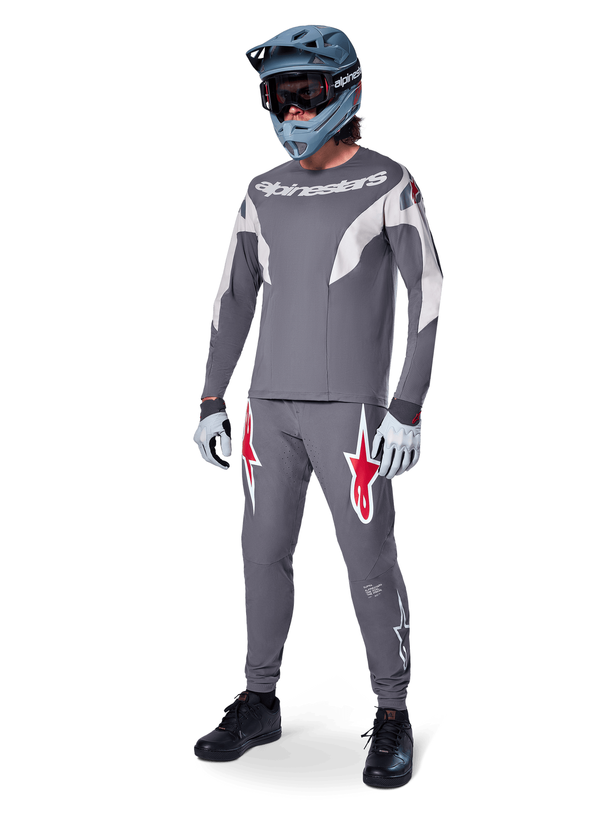 Pantalones Alpinestars A-Supra Race en gris oscuro, con logotipos llamativos en rojo y blanco en los muslos, usados por un piloto como parte de un kit completo de carreras de MTB con jersey gris a juego, casco integral gris azulado y guantes gris claro, equipamiento de alto rendimiento para ciclismo de montaña