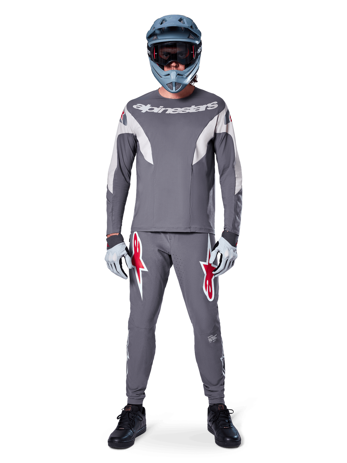 Ciclista de MTB vistiendo pantalones Alpinestars A-Supra Race en gris oscuro, con llamativos logotipos rojos en los muslos, combinados con un jersey gris a juego, casco integral azul y zapatillas negras para un equipamiento completo de competición off-road