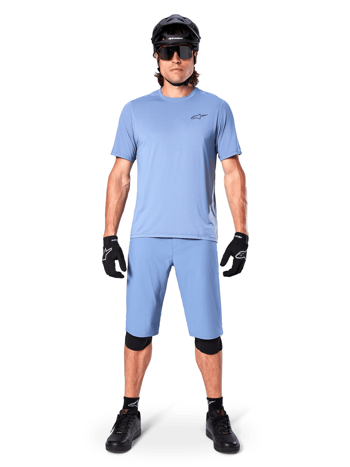 Modelo con los shorts Alpinestars A-Dura en color Infinity Blue, jersey azul claro a juego, casco de bicicleta de montaña negro, gafas de sol negras, guantes y rodilleras negros, equipamiento completo de MTB para trail