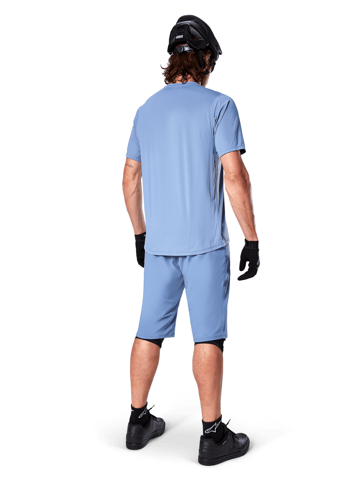 Vista trasera de un ciclista de montaña con shorts Alpinestars A-Dura en color Infinity Blue, jersey de manga corta a juego, casco negro, guantes negros y zapatillas negras, equipamiento coordinado de alto rendimiento para trail MTB