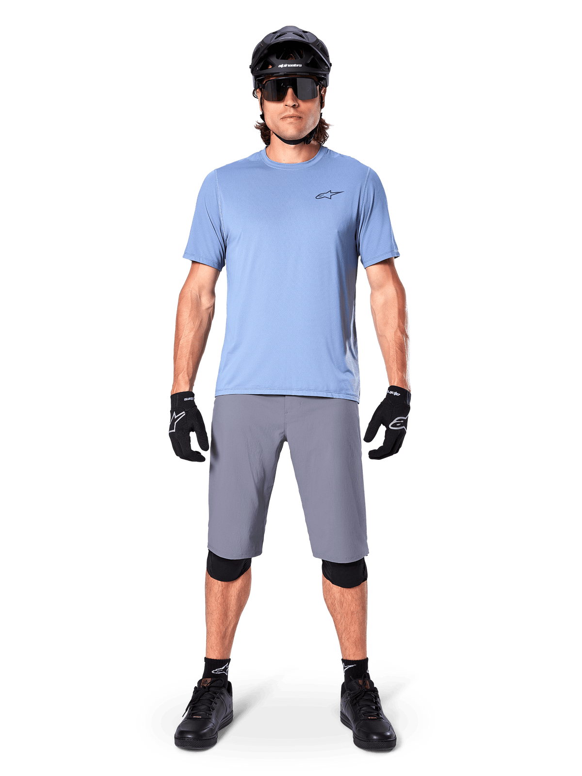 Ciclista masculino con Alpinestars A-Dura Shorts en gris oscuro, combinados con una camiseta de manga corta azul claro, casco negro, gafas de sol y rodilleras negras, equipamiento completo de ciclismo de montaña para rendimiento off-road
