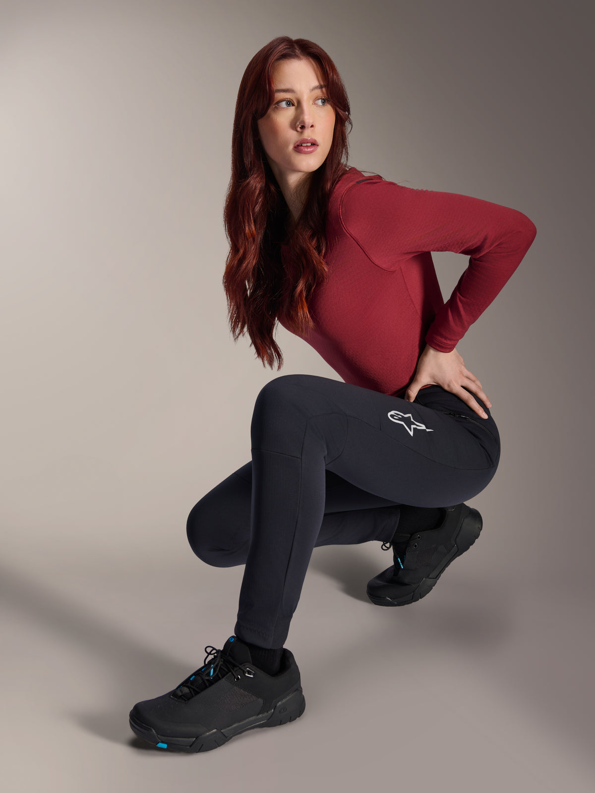 Alpinestars Stella A-Dura Thermal Pants, pantalones de MTB para mujer, negros, usados por una modelo en una pose agachada con una camiseta de manga larga granate y zapatillas de ciclismo negras, con el logotipo de la estrella blanca en el muslo, construcción duradera y cálida para ciclismo todoterreno en climas fríos