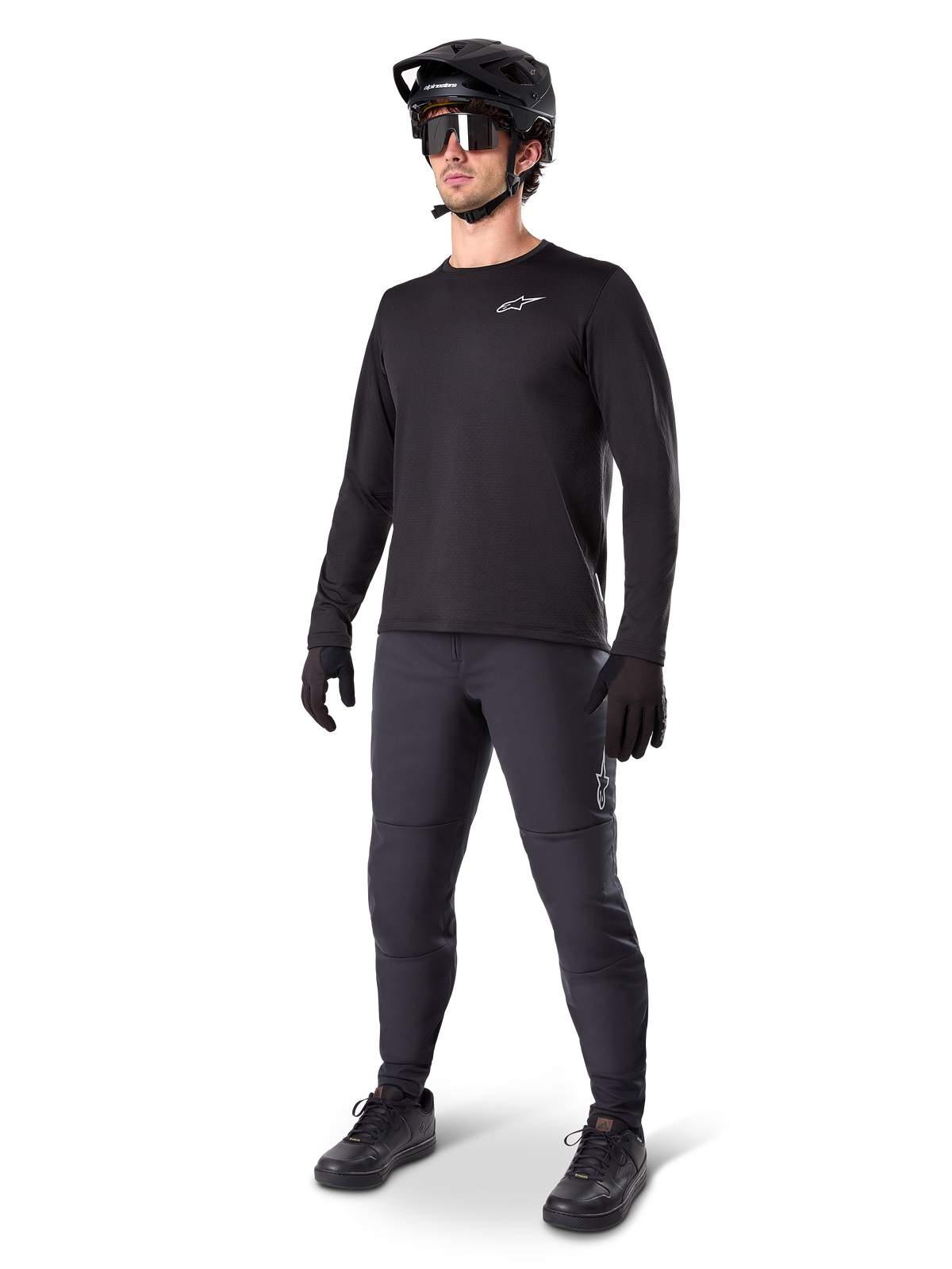 Modelo con el Jersey Térmico Alpinestars A-Dura, jersey de MTB de manga larga negro, casco de MTB negro, gafas de sol y pantalones gris oscuro, con un pequeño logotipo blanco en el pecho y forro polar aislante para montar en climas fríos.